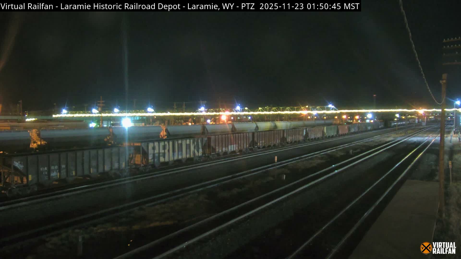 The image shows a nighttime view of a railroad yard with multiple tracks and several long freight trains under a clear sky, illuminated by numerous bright lights.