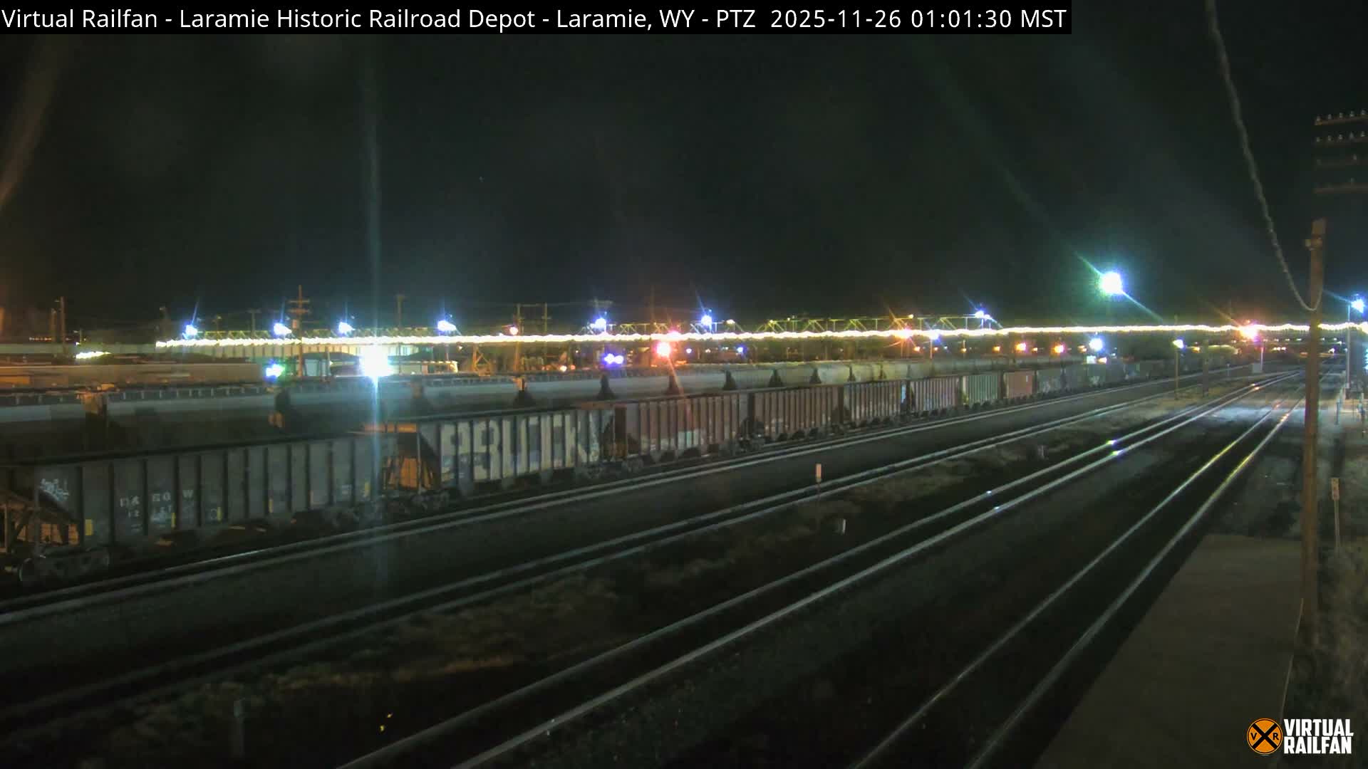 A clear night view shows multiple sets of dark train tracks in the foreground and a long freight train illuminated by bright lights in a distant rail yard or industrial area under a dark sky.