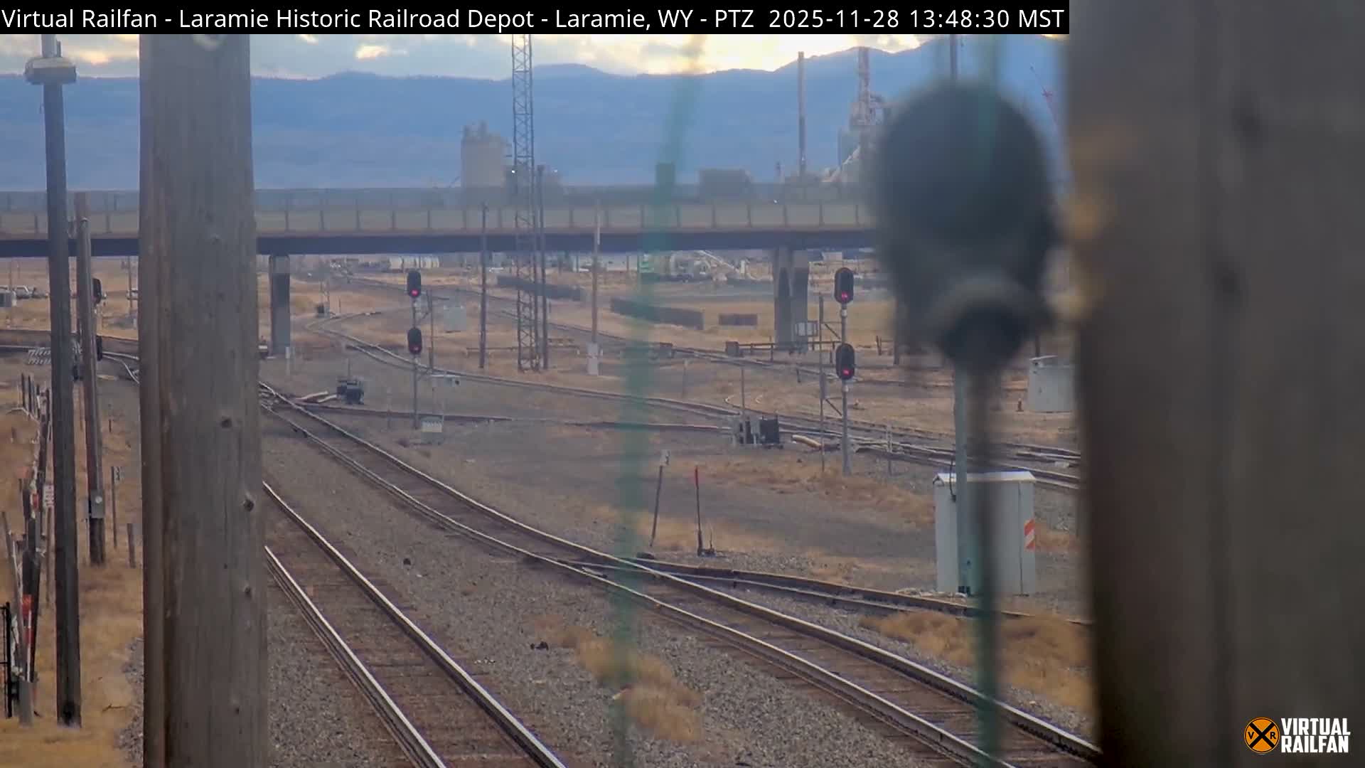 Under an overcast sky, a wide view shows an extensive railroad switching yard with multiple tracks, active red signals, an overhead bridge, and industrial buildings against distant mountains.