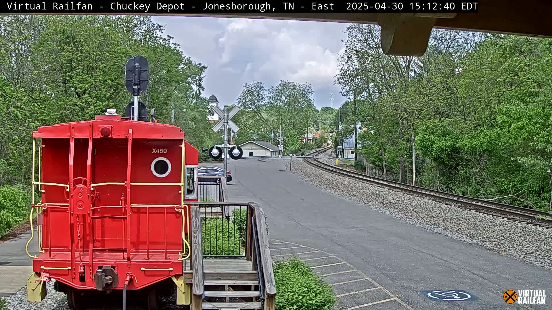 A red railroad caboose sits in front of a railway track that curves to the right, under a partly cloudy sky.