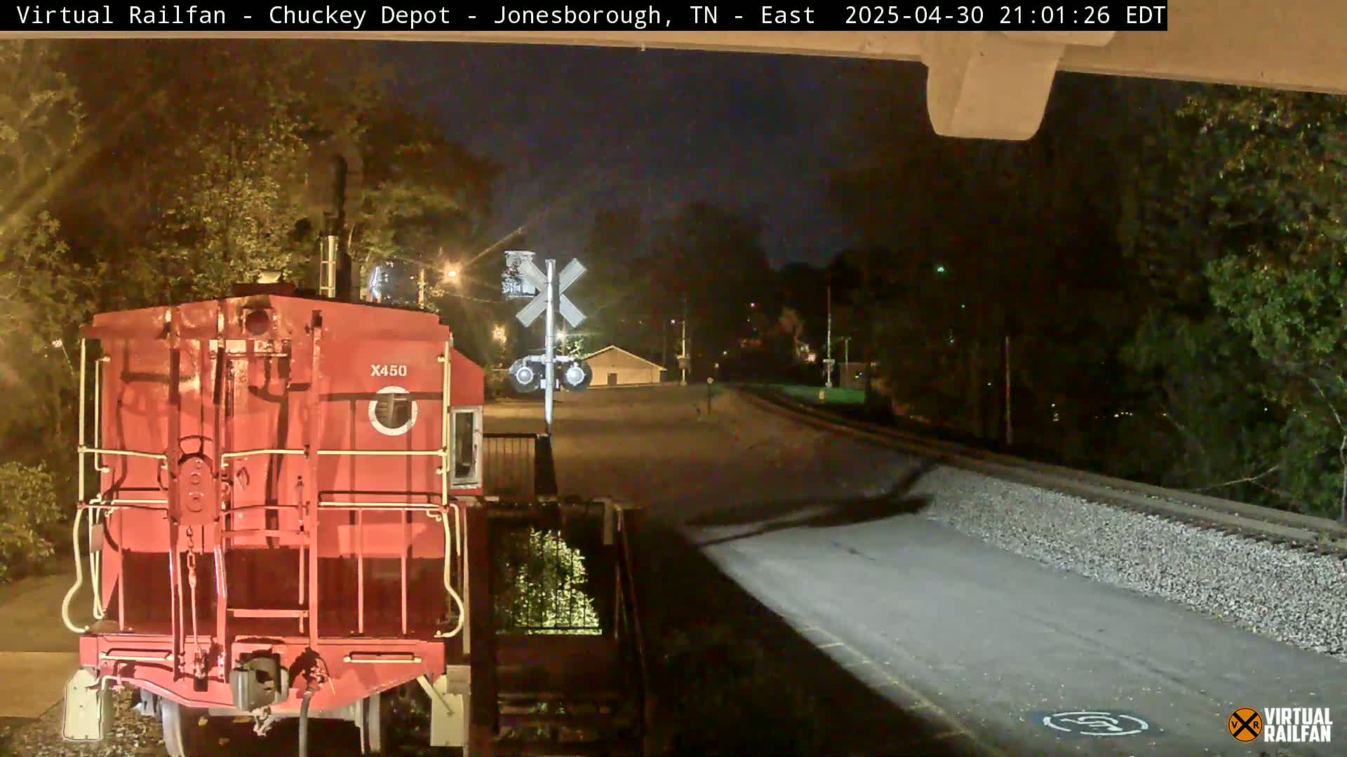 A red train car is stopped at a nighttime railroad crossing near some trees under a dark sky.