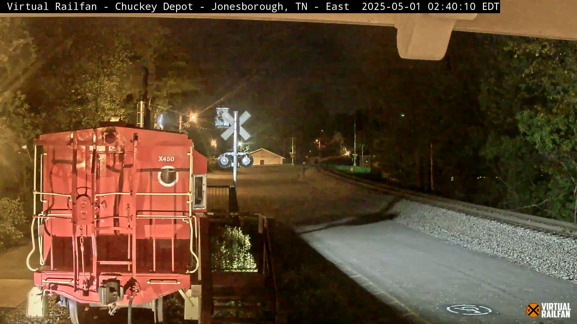 At night, a red train car is visible at a railroad crossing near a road, with trees and a small building in the background.