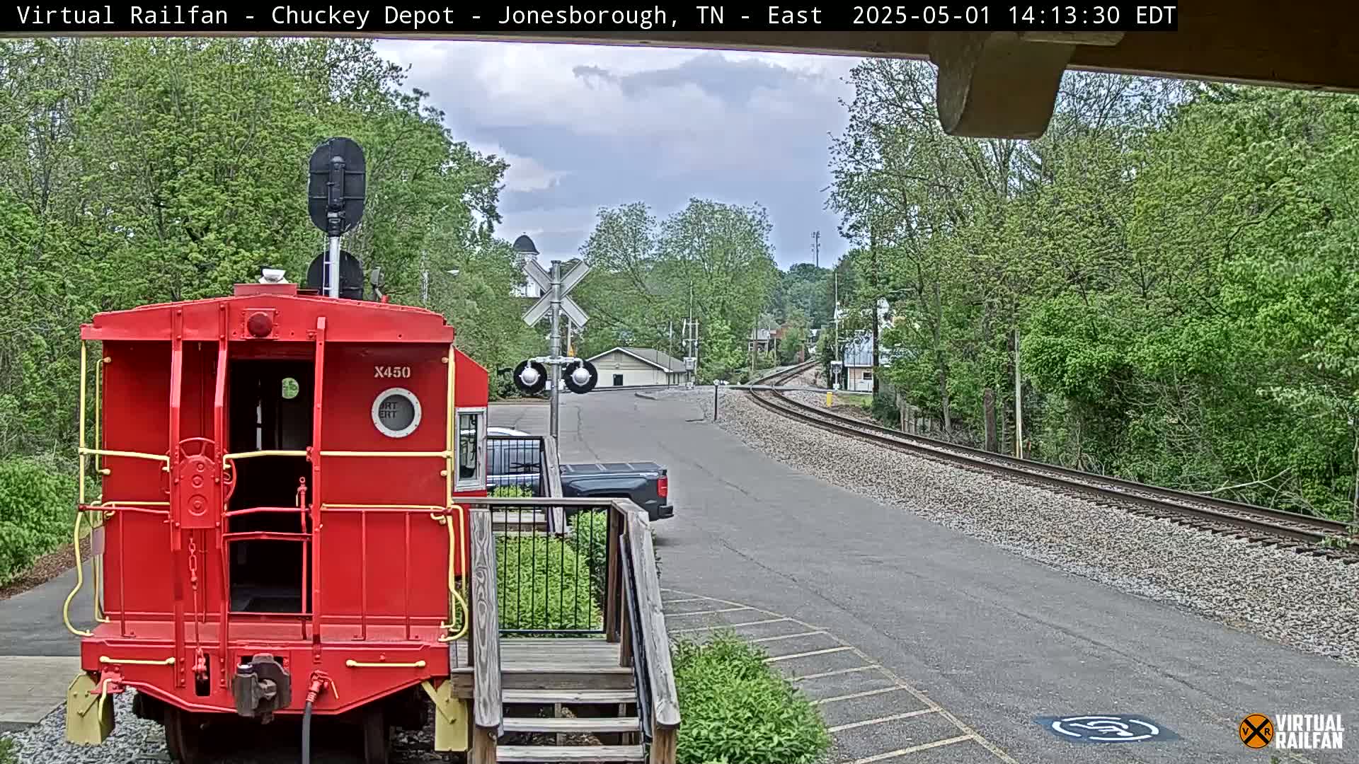 A red caboose sits in the foreground, facing away from a train track that curves to the right, under a partly cloudy sky, with green trees lining the track and a paved area in the immediate vicinity.