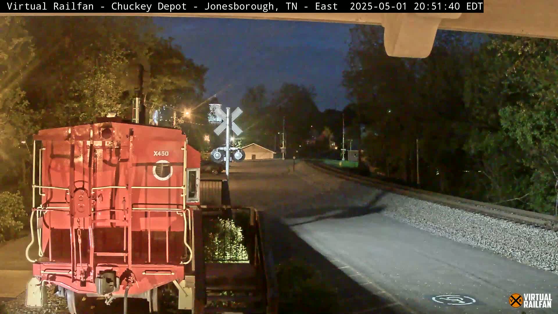 Here is a description of the image: A red train engine is parked at a nighttime railroad crossing, under a dark sky, near trees and a small building.