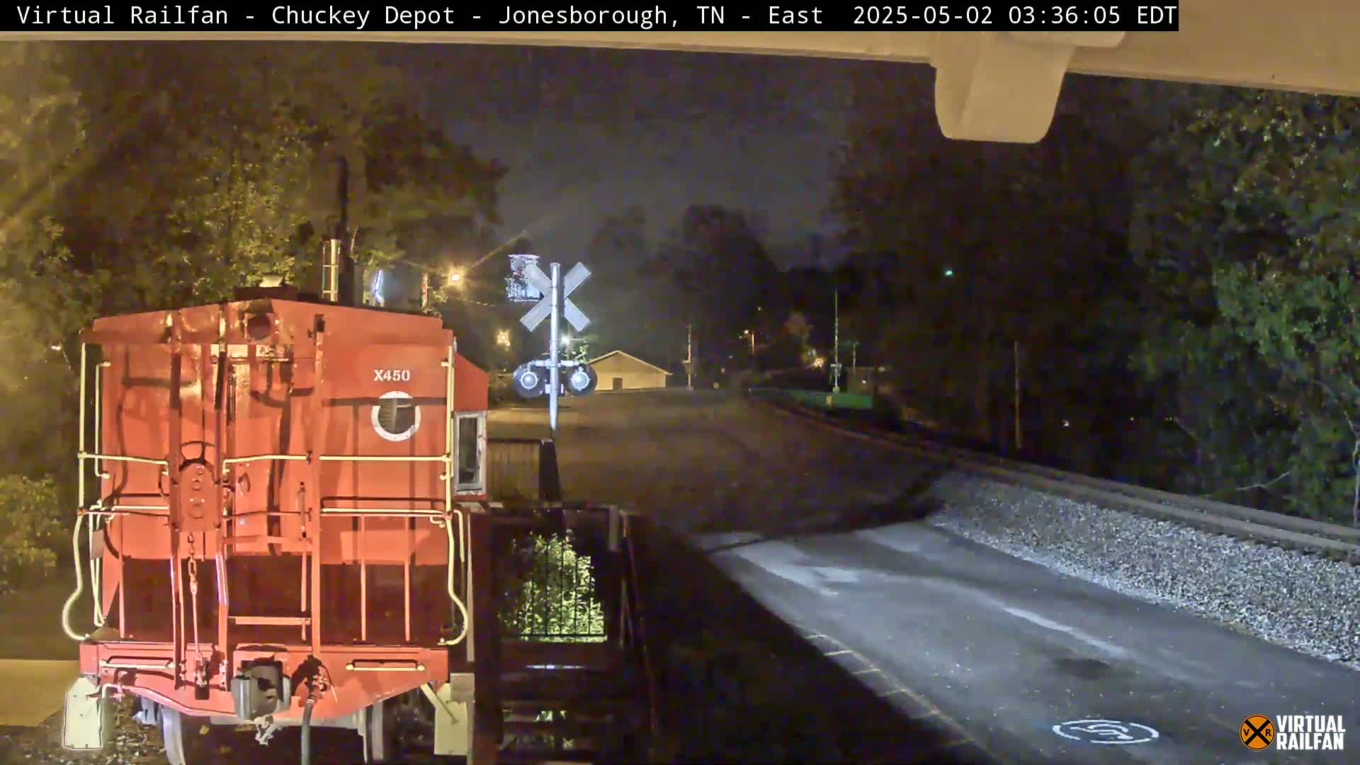 At night, a red train car sits at a railroad crossing near a roadway and some trees.