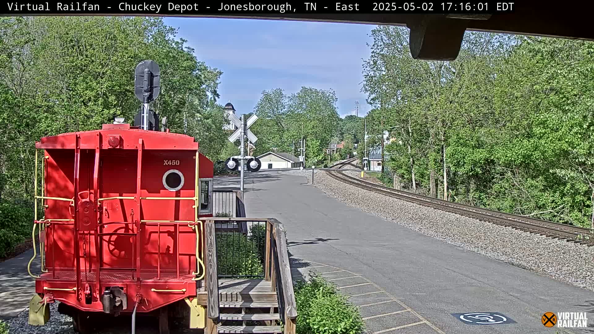 A red train car sits in front of a railroad crossing on a sunny day, with a view of train tracks and leafy green trees in the background.