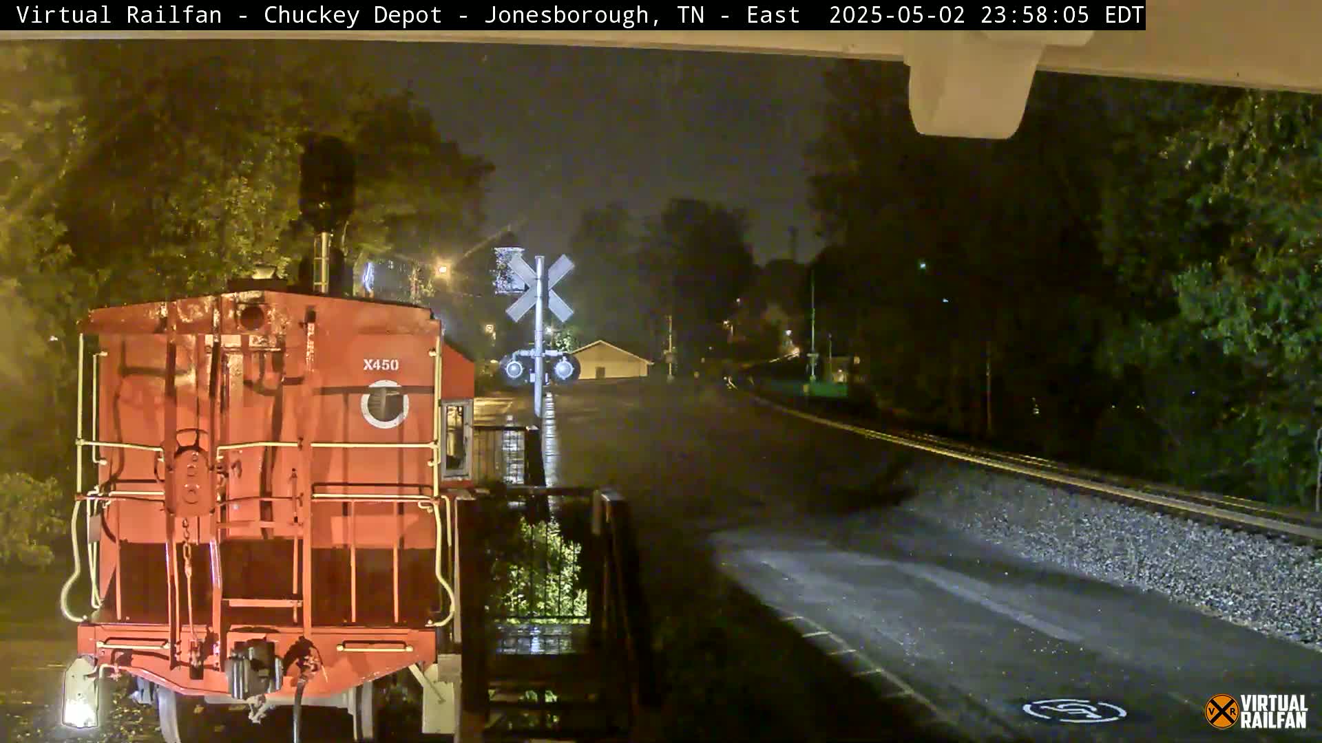 A red railroad car is stopped at a nighttime railway crossing under rainy conditions.