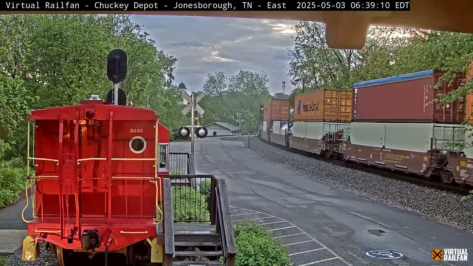 A red railcar sits in the foreground, facing a freight train passing by under an overcast sky.
