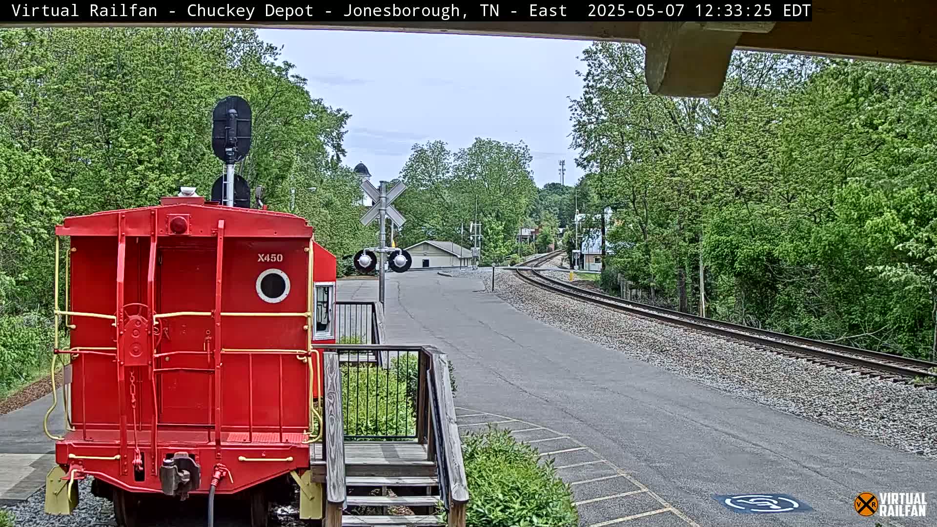 A red railroad car is parked at a railway crossing on a partly cloudy day, with lush green trees lining the tracks and a paved road nearby.