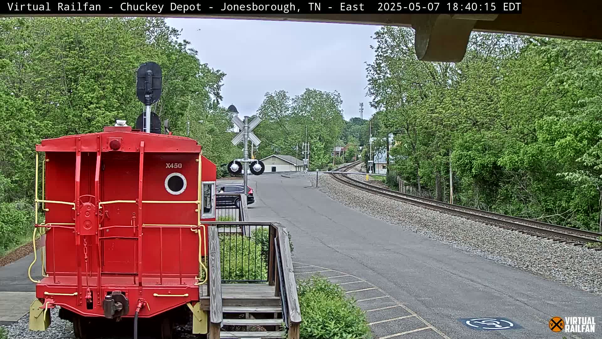 A red railroad car is parked next to a roadway and railroad tracks, surrounded by lush green trees on a partly cloudy day.