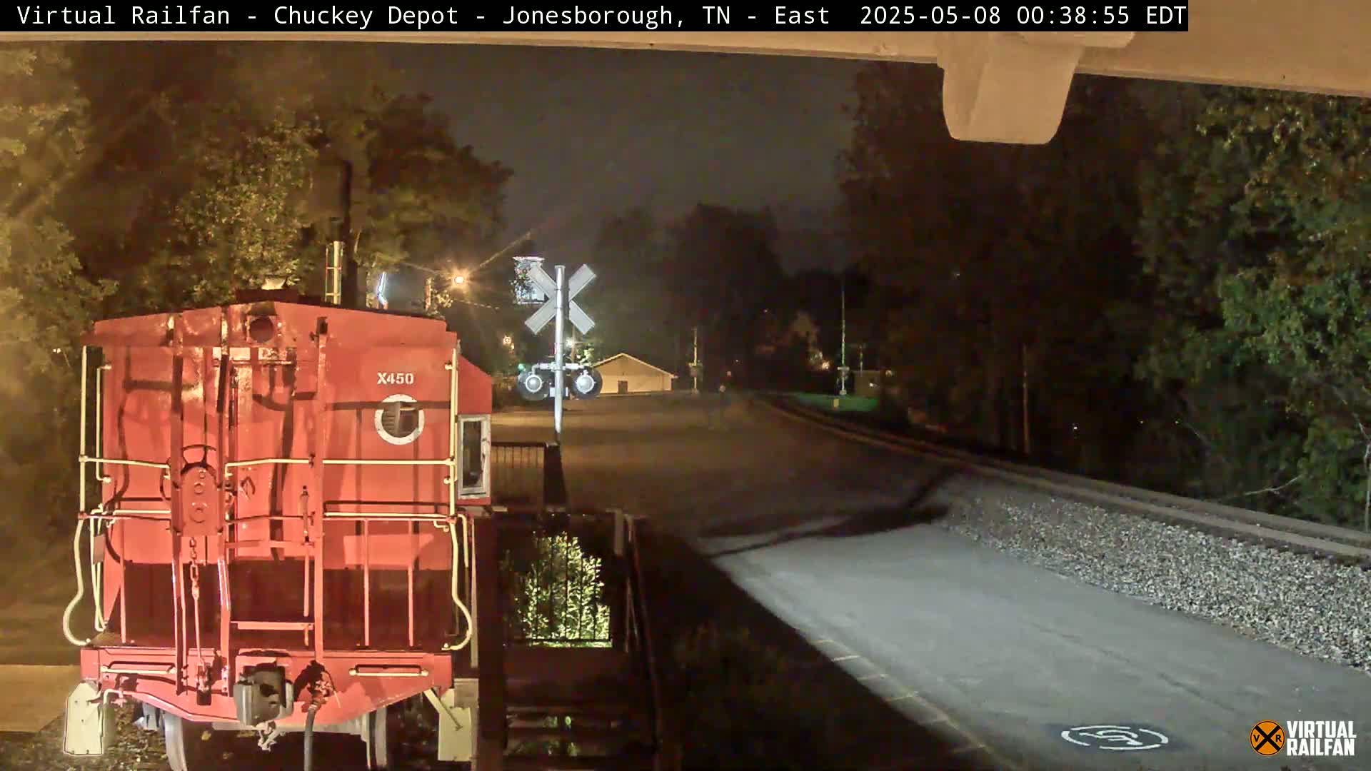 A red train car sits at a nighttime railroad crossing near some trees and a building under a clear sky.