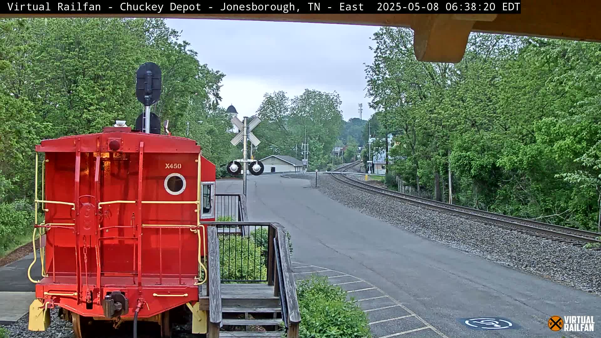 A red railway car is parked next to a railroad crossing on an overcast day, with a road leading to a treeline in the background.