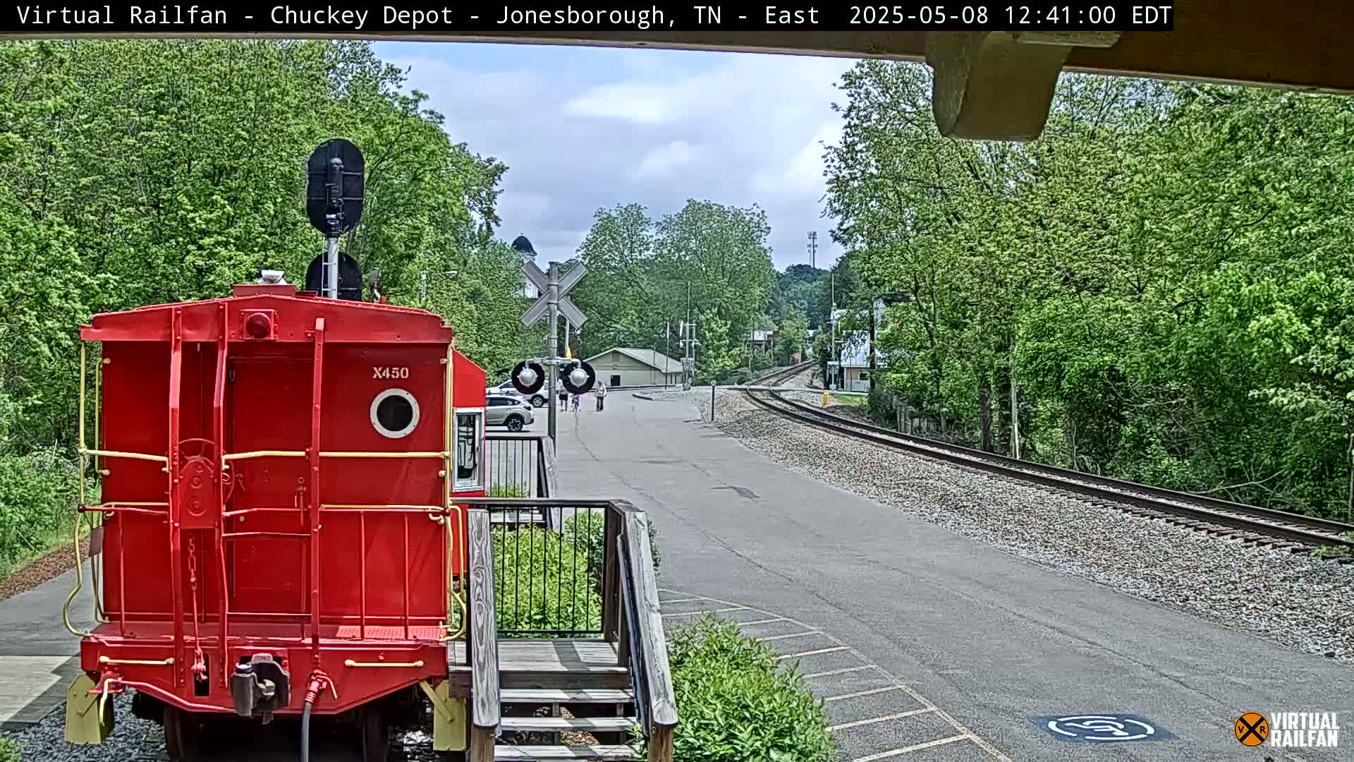 A red railroad car sits on a track next to a paved road, with a rail crossing and lush green trees in the background under a partly cloudy sky.