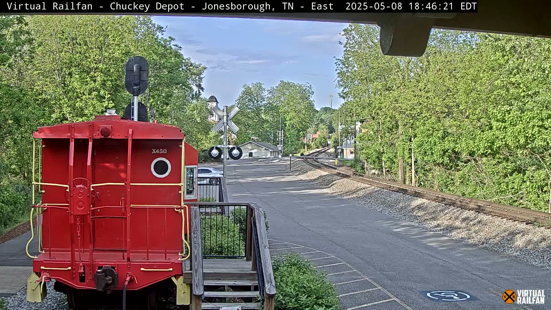A red railway car is parked near a railway line, with a partly sunny sky and lush green trees in the background.
