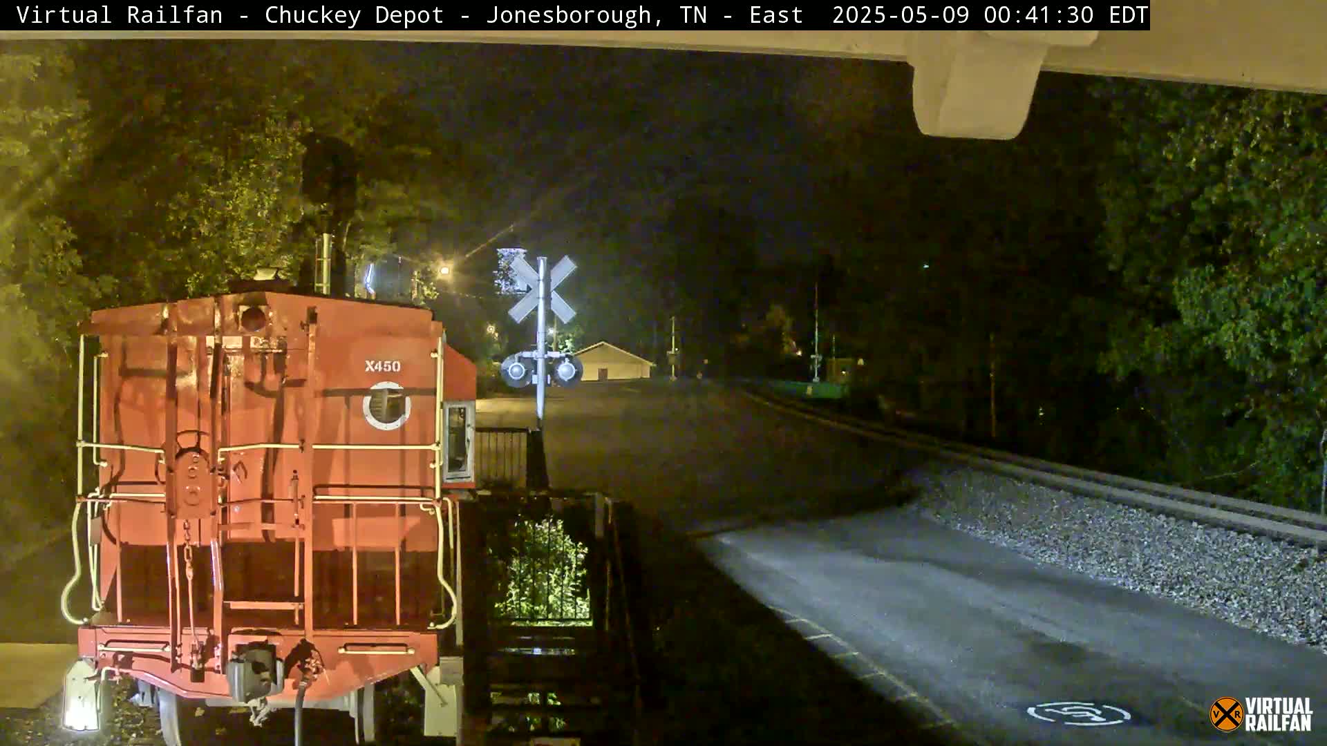 At night, a red railroad car sits at a railroad crossing near a road, under the cover of trees.