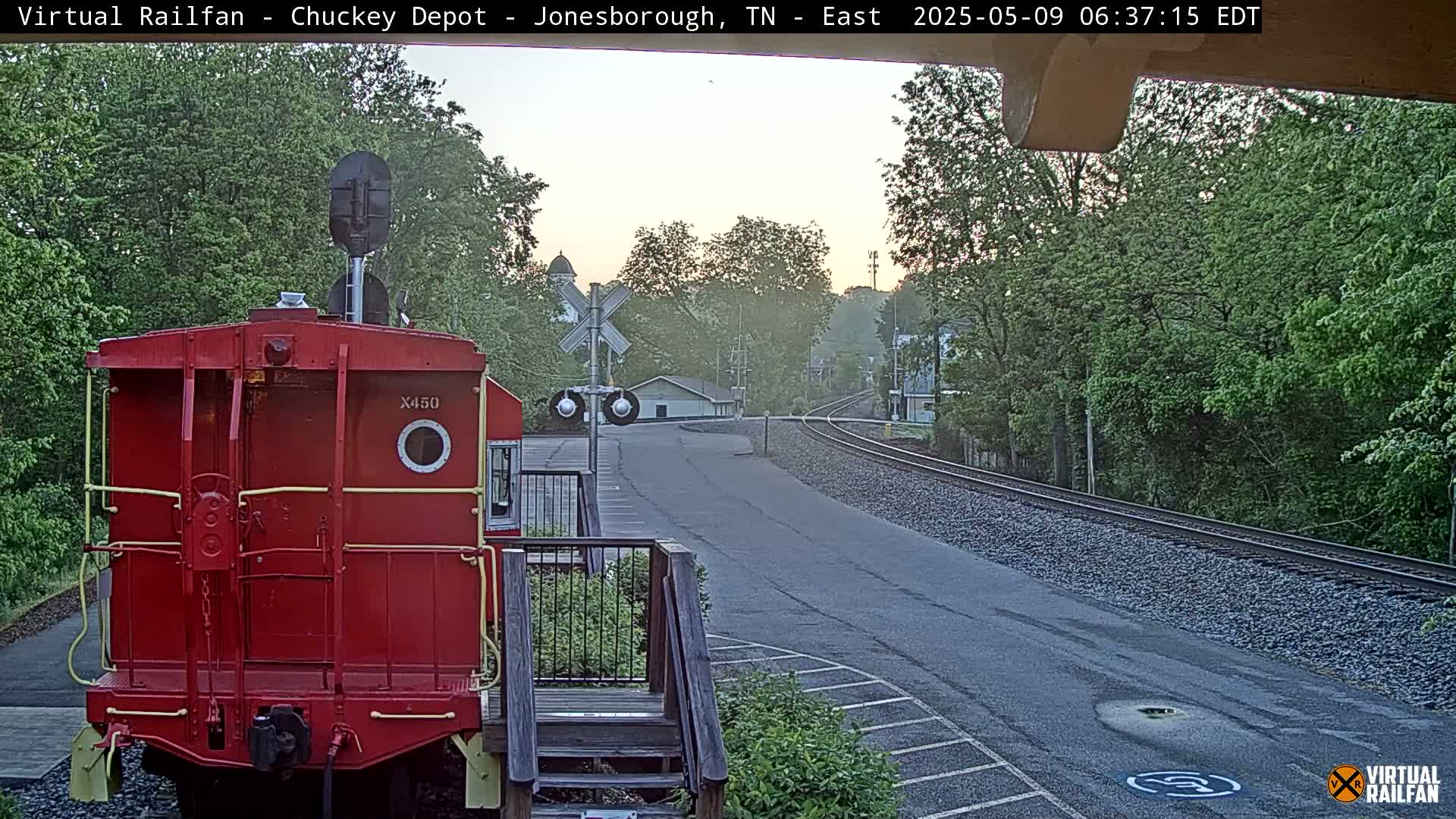 A red railway car faces the viewer, situated near railway tracks that extend into a town under a hazy, dawn sky.
