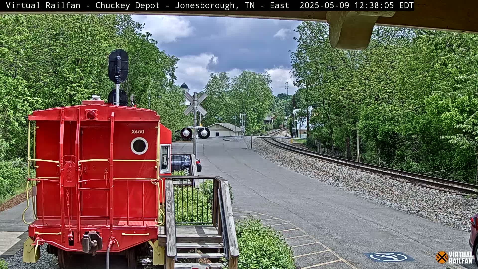A red railroad car is parked near a train track that curves into a leafy, partly cloudy landscape.