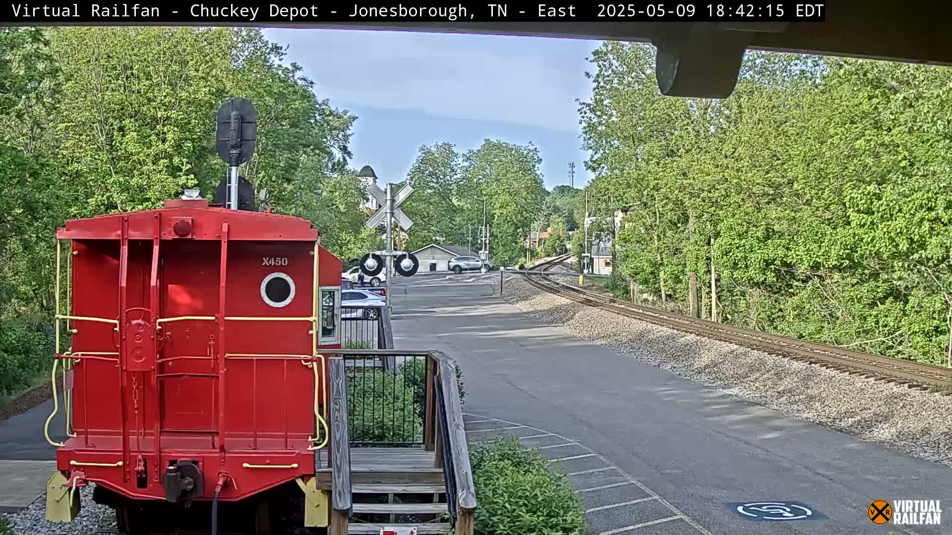 A red railroad car is parked near a railway crossing with a road and several vehicles visible under a partly sunny sky.