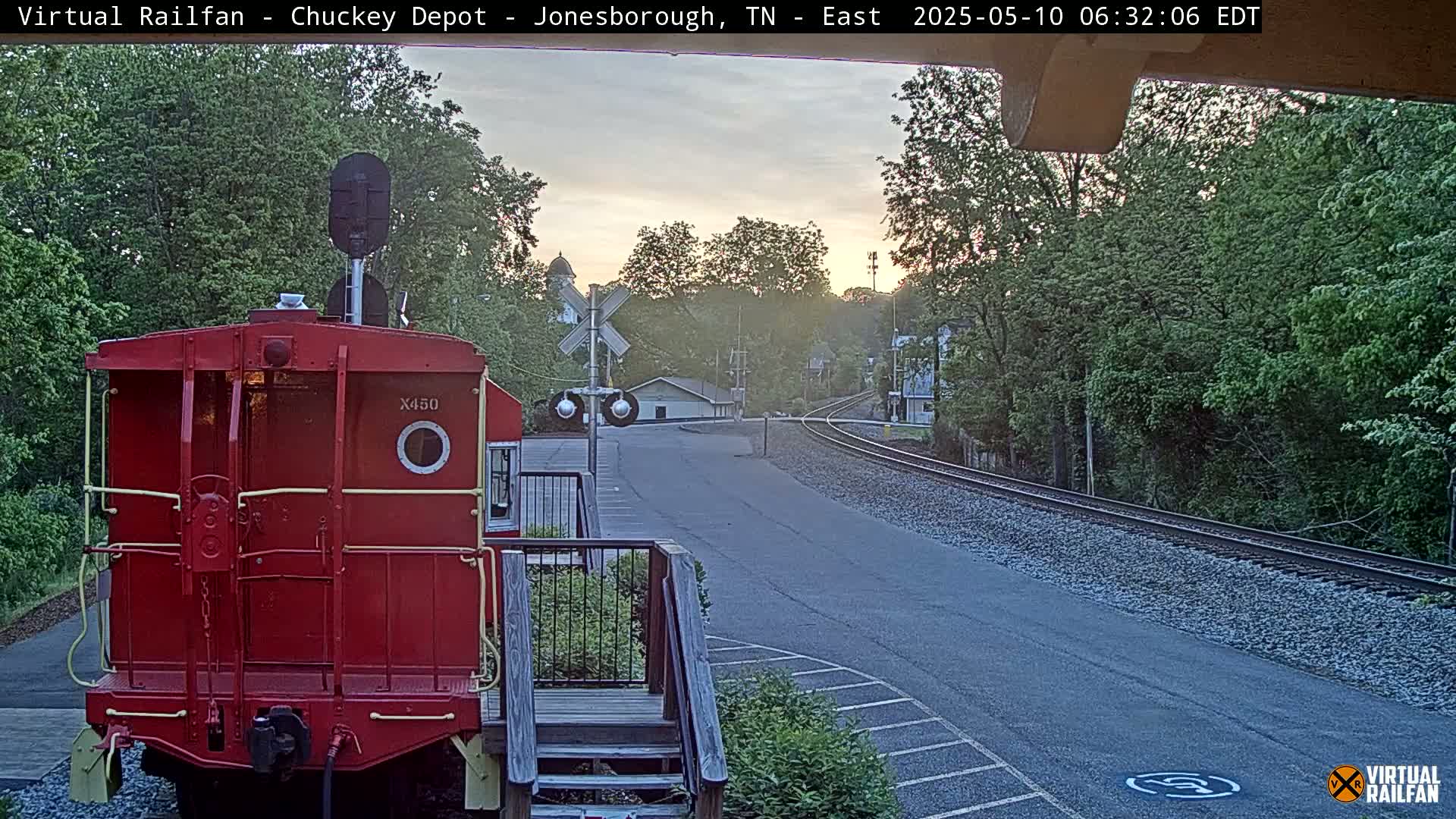 A red train car is parked at a train depot at sunrise, with railroad tracks extending into a tree-lined area.