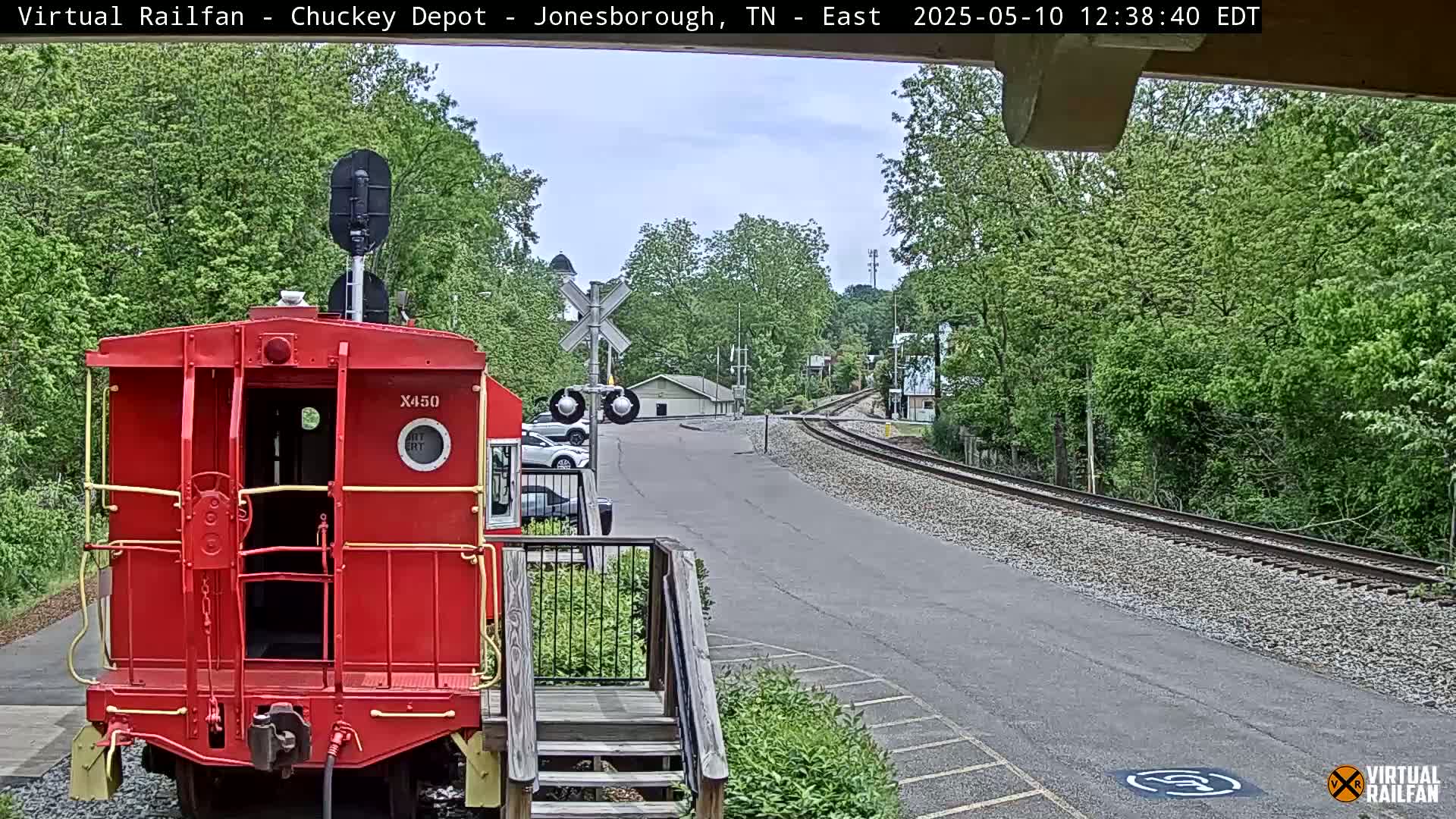 A red railway caboose sits in front of a paved road and railroad tracks, surrounded by lush green trees under a partly cloudy sky.