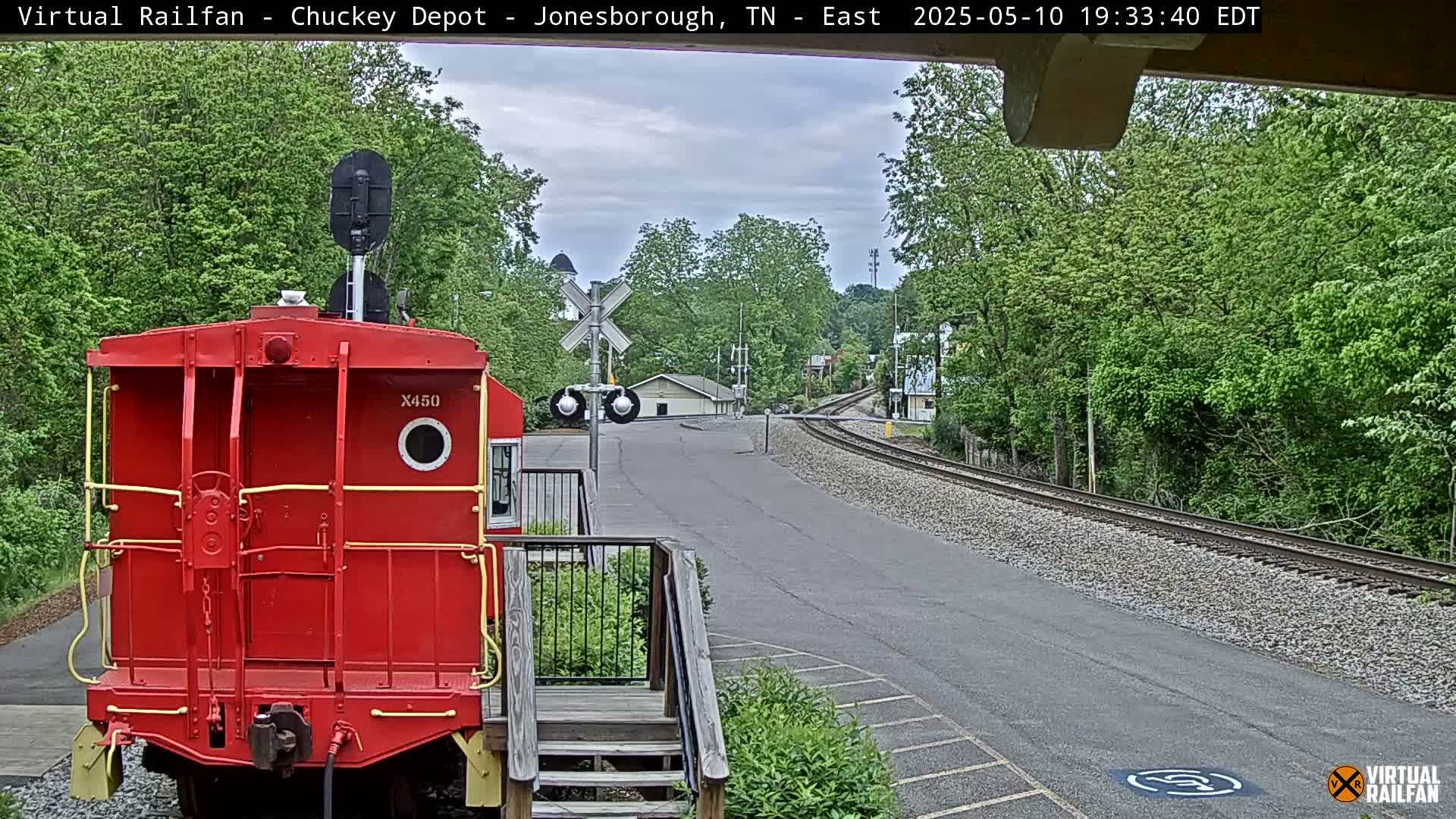 A red caboose sits at a train depot overlooking a railway track that curves into a tree-lined town under a partly cloudy sky.