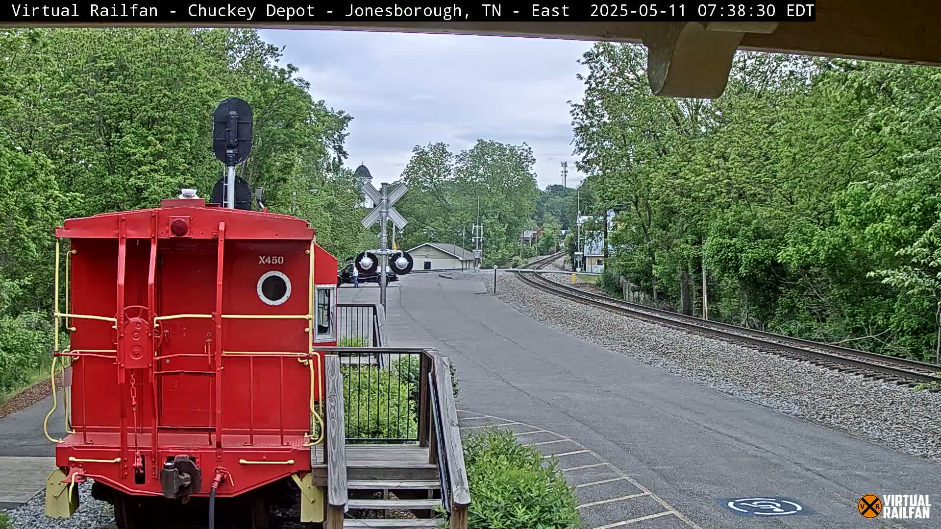 A red train caboose is parked at a railroad crossing on an overcast day, with lush green trees lining the tracks and a paved road nearby.