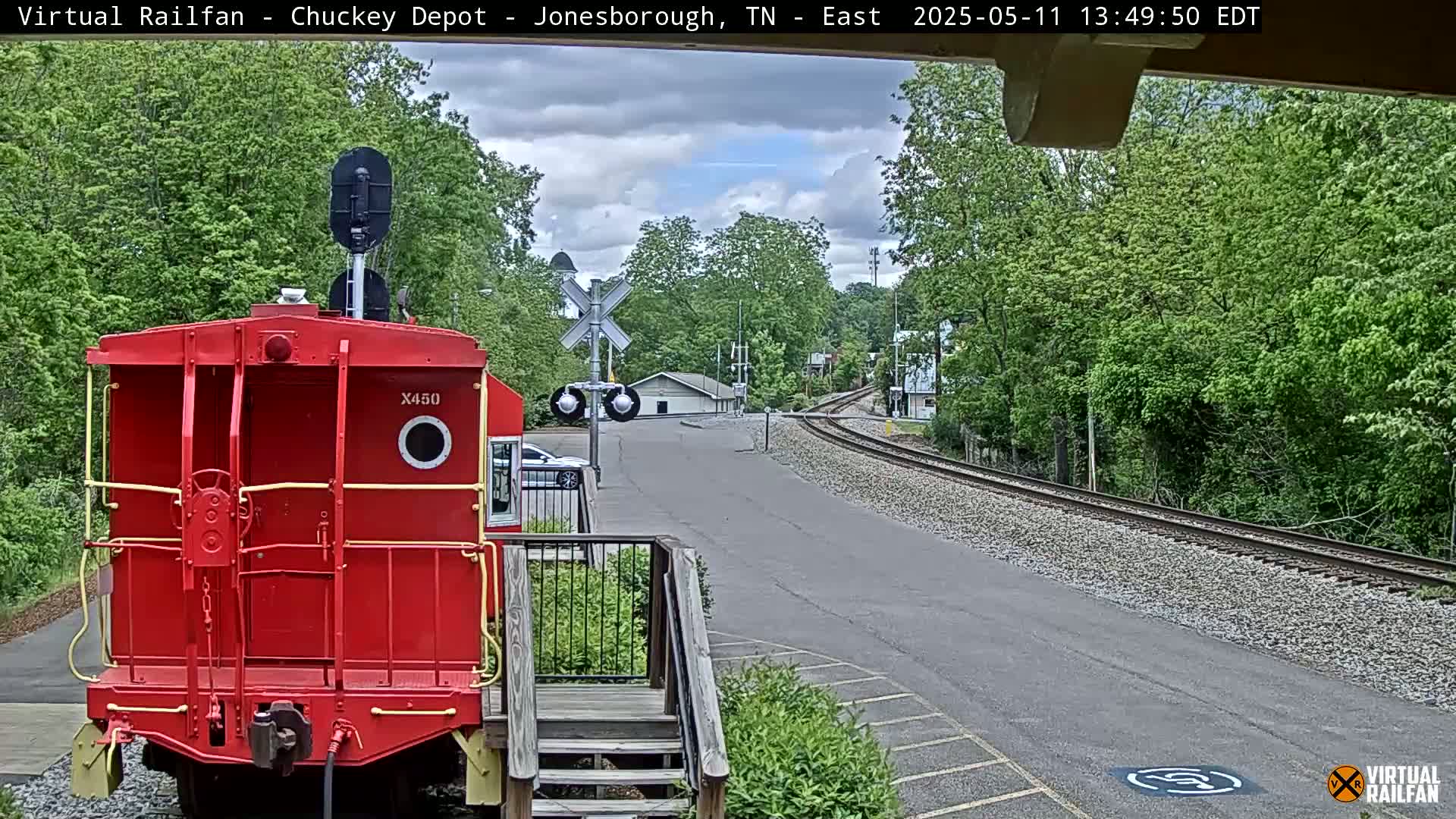 A red railroad car is parked facing away from the camera at a train depot on a partly cloudy day, with railroad tracks curving away into a wooded area.