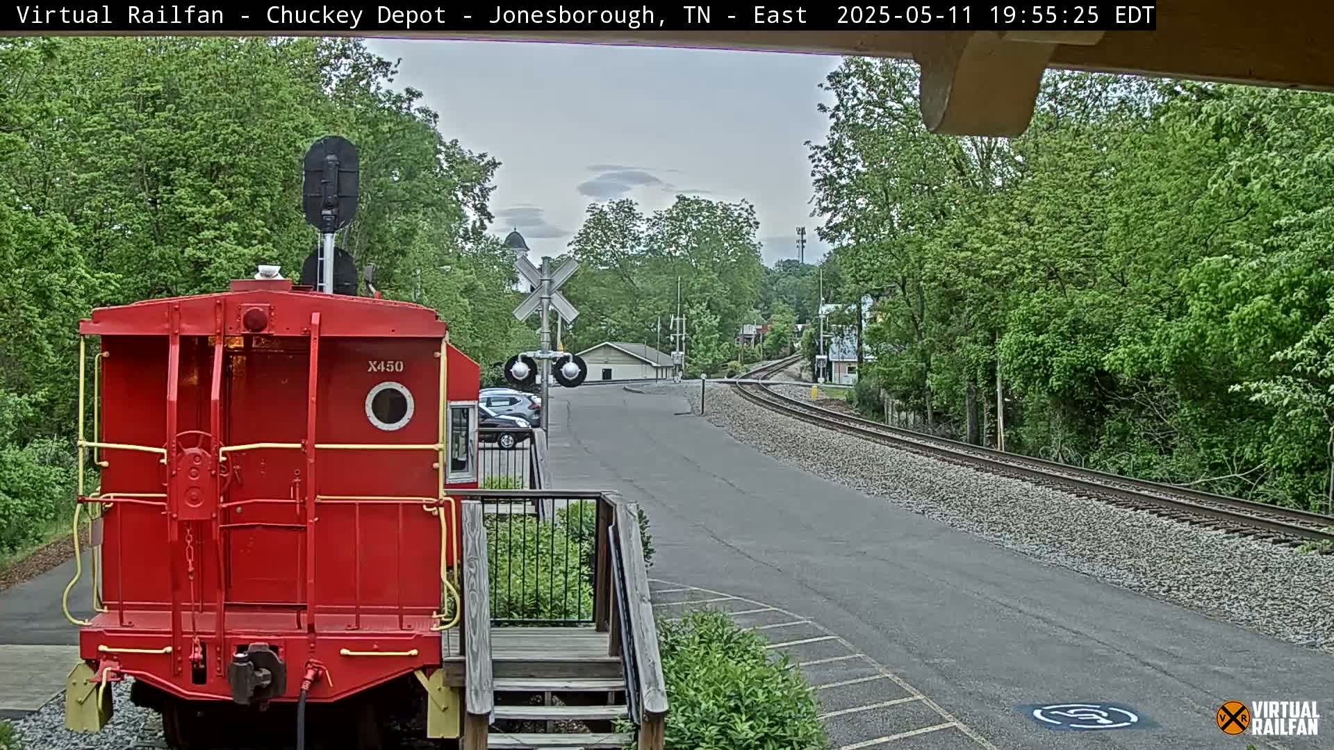 A red train car is parked at a depot overlooking a curving railway track under an overcast sky, surrounded by lush green trees.