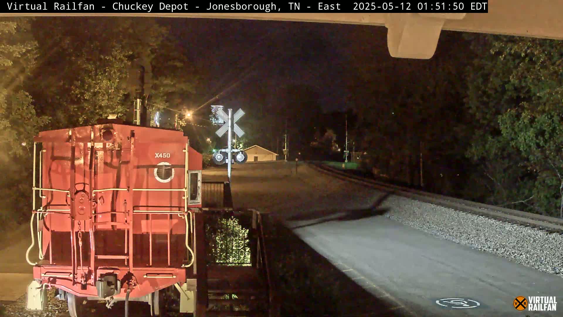 A red train car is stopped at night at a railroad crossing near some trees.