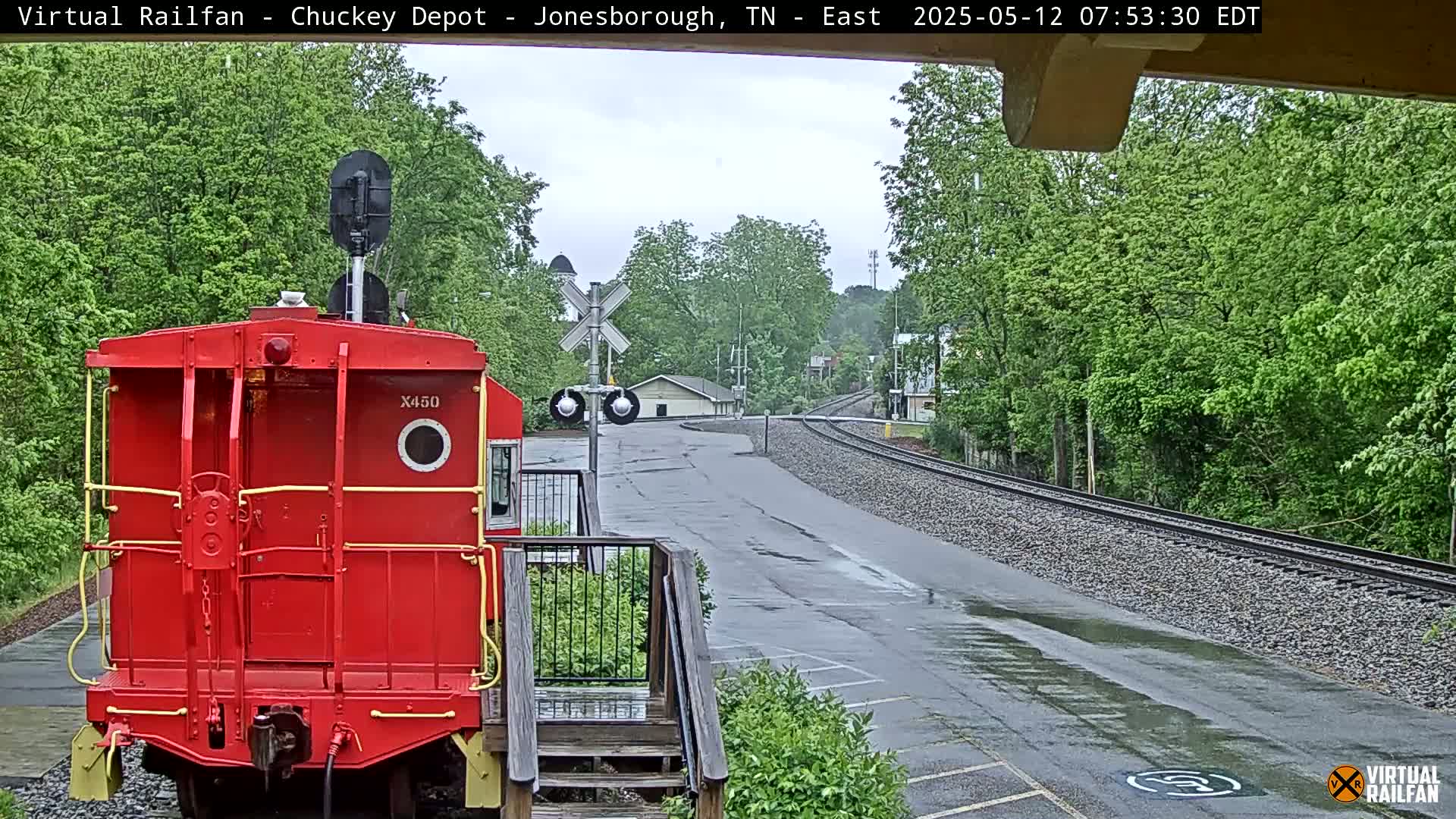 A red train car is parked near a railway track on a wet, overcast day, with lush green trees and a railroad crossing visible in the background.