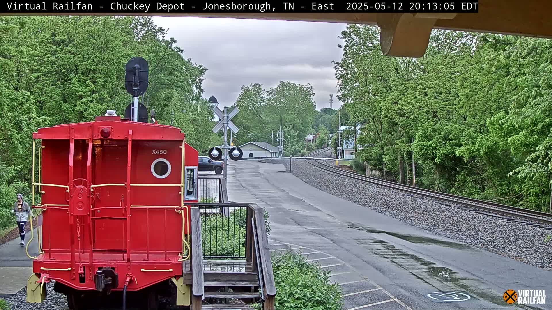 A red train caboose is parked next to a road and railway tracks, with a person walking in the background under an overcast sky.