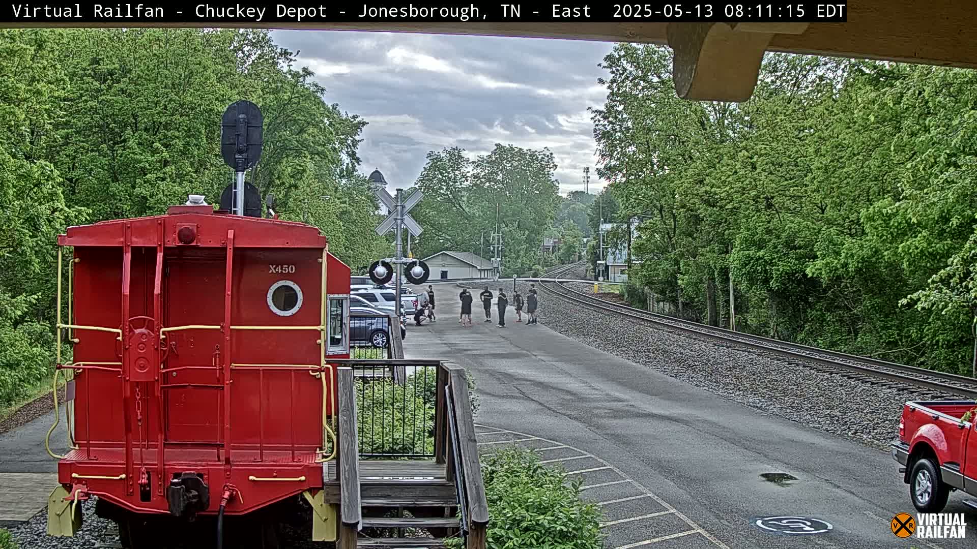 A red train car is parked in front of a railroad crossing where a group of people stand, all under a partly cloudy sky.