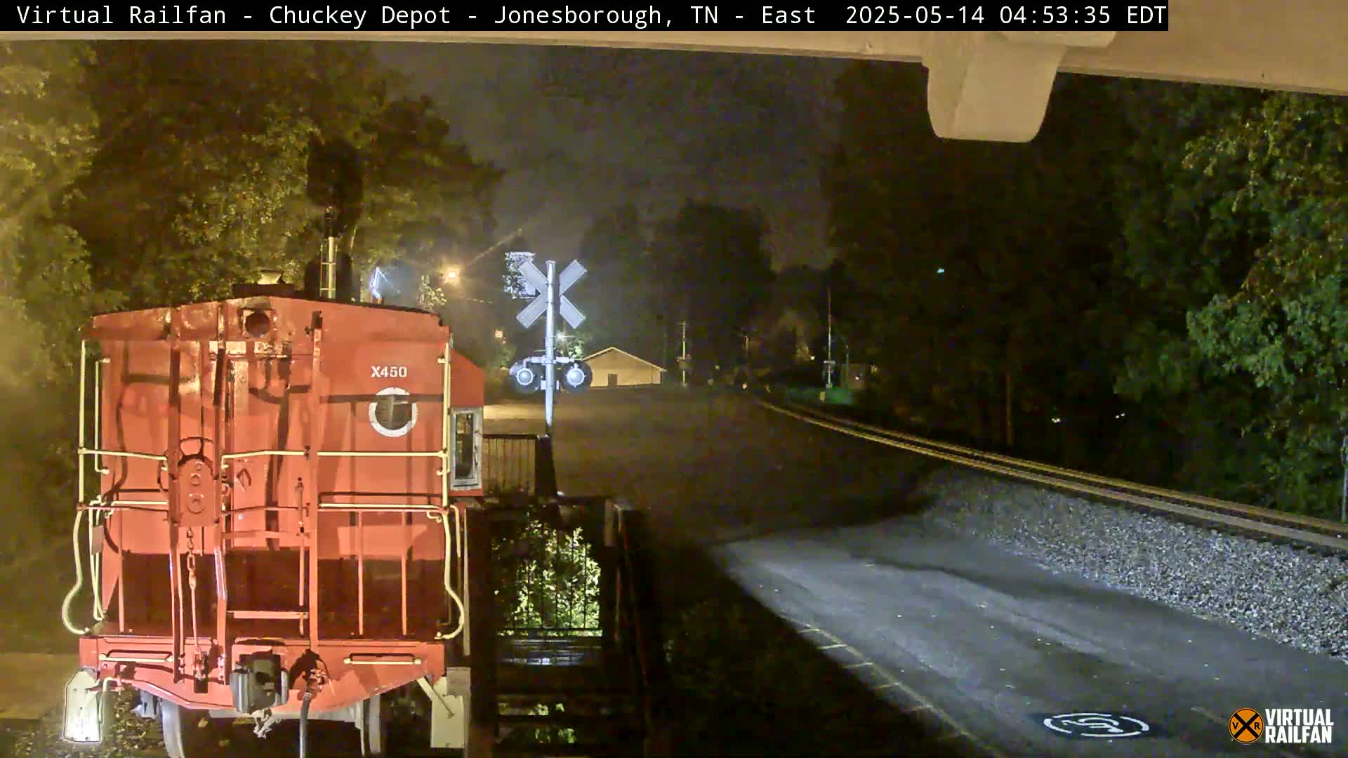 A red train car is stopped at night at a railroad crossing near trees and a building.