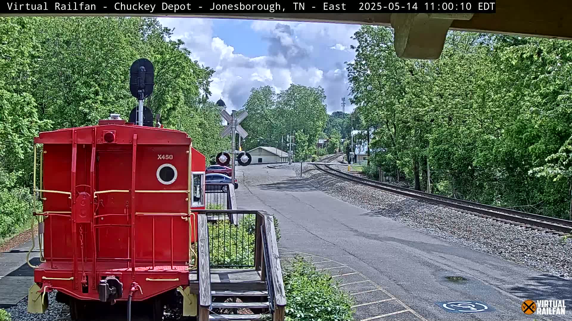 A red train car is parked in front of a railroad crossing on a partly cloudy day, with lush green trees lining the tracks.