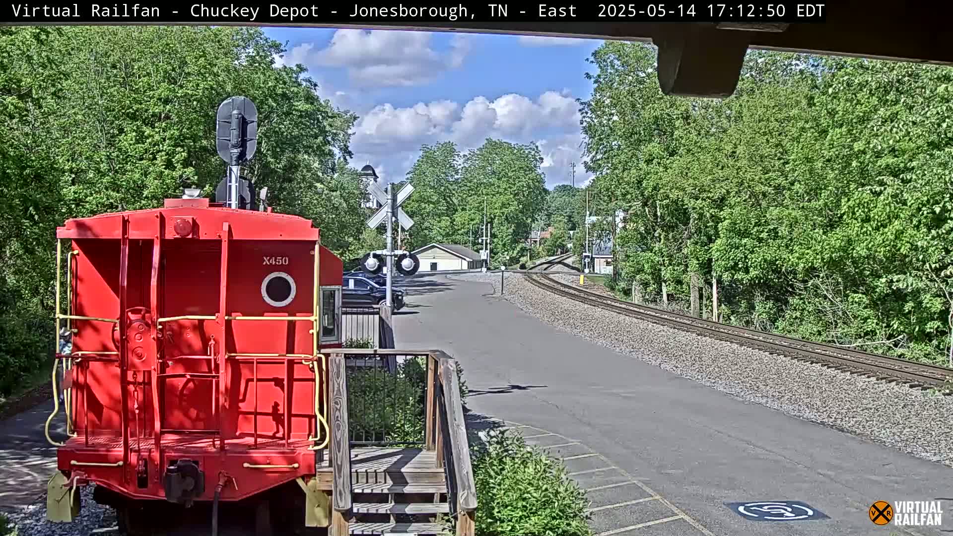 A red train car is parked at a train depot on a partly sunny day, with a rail line extending into the distance.