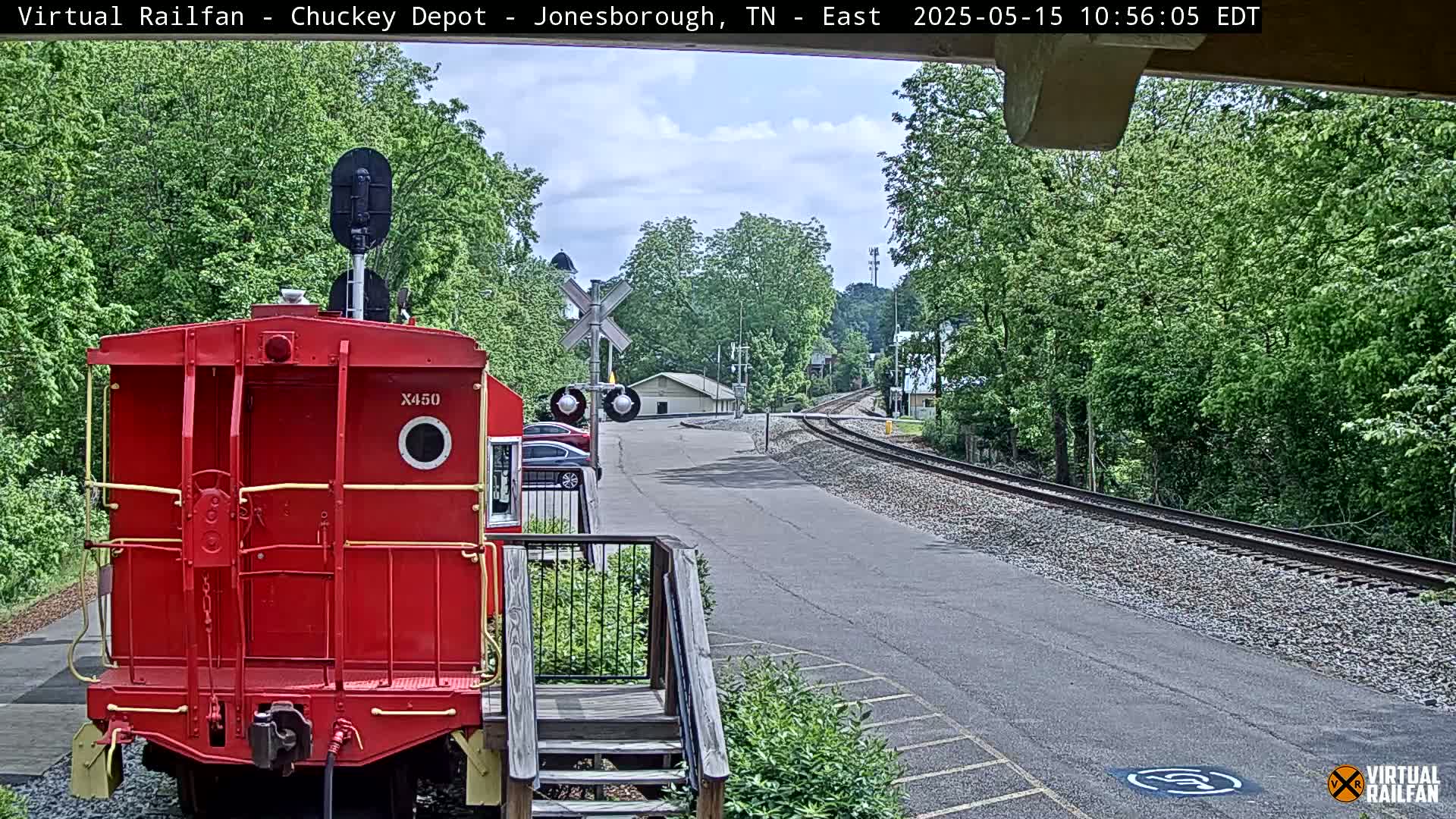A red railroad car sits in the foreground, facing away from a train track that curves away into a tree-lined area under a partly cloudy sky.
