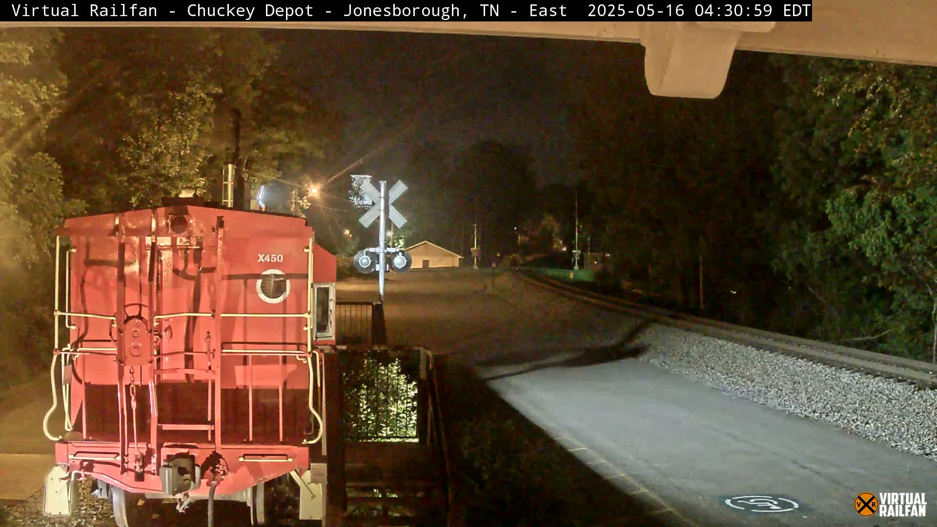At night, a red train car sits at a railroad crossing near trees and a small building.