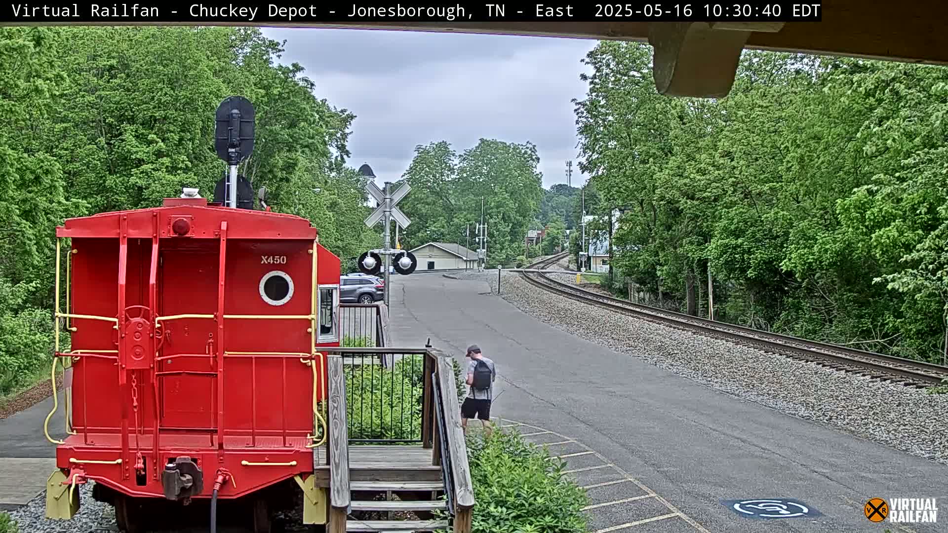 A red train car is parked at a train station on a partly cloudy day, with a person walking nearby.