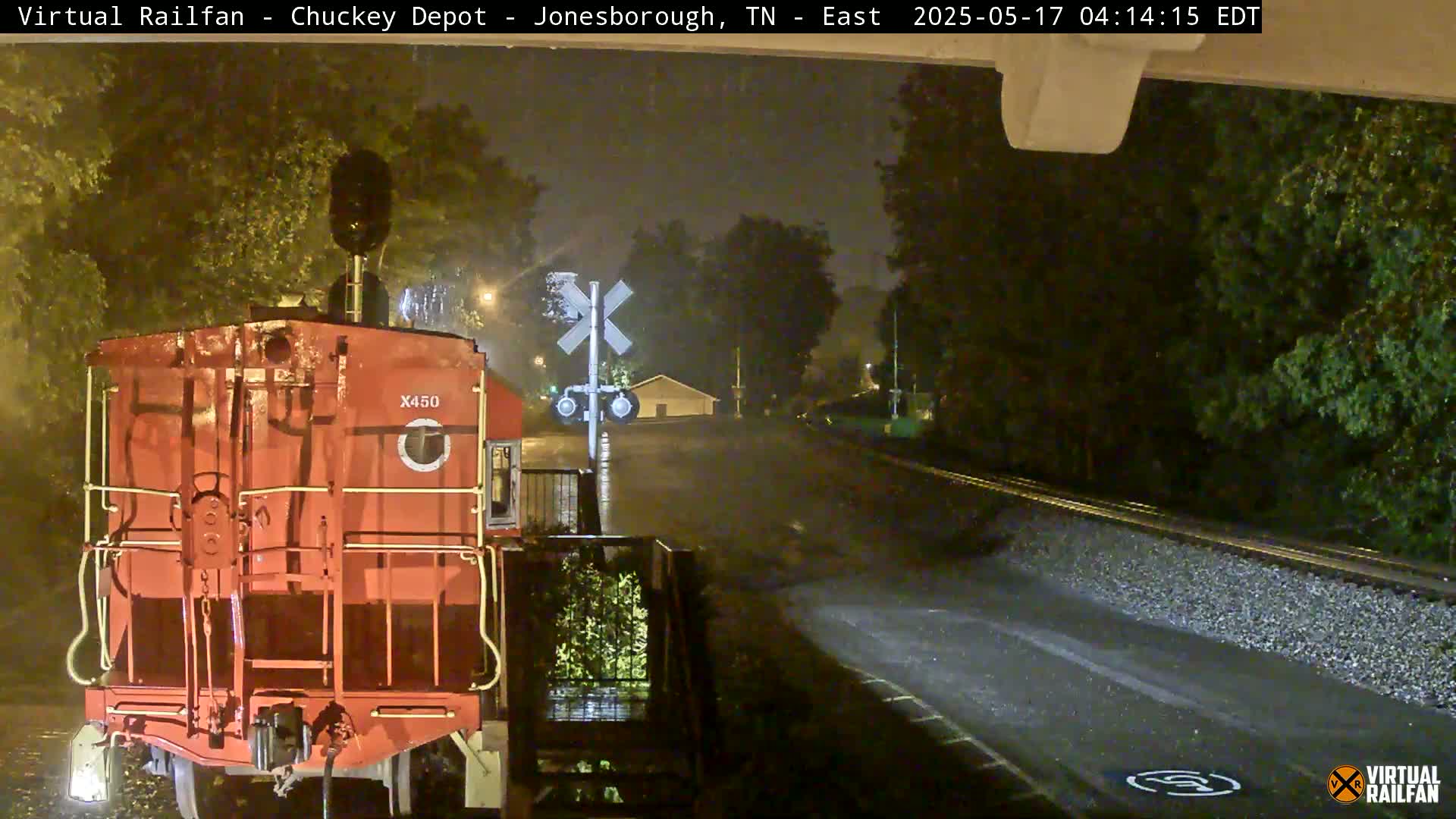 A red train car sits at a railroad crossing at night in the rain.