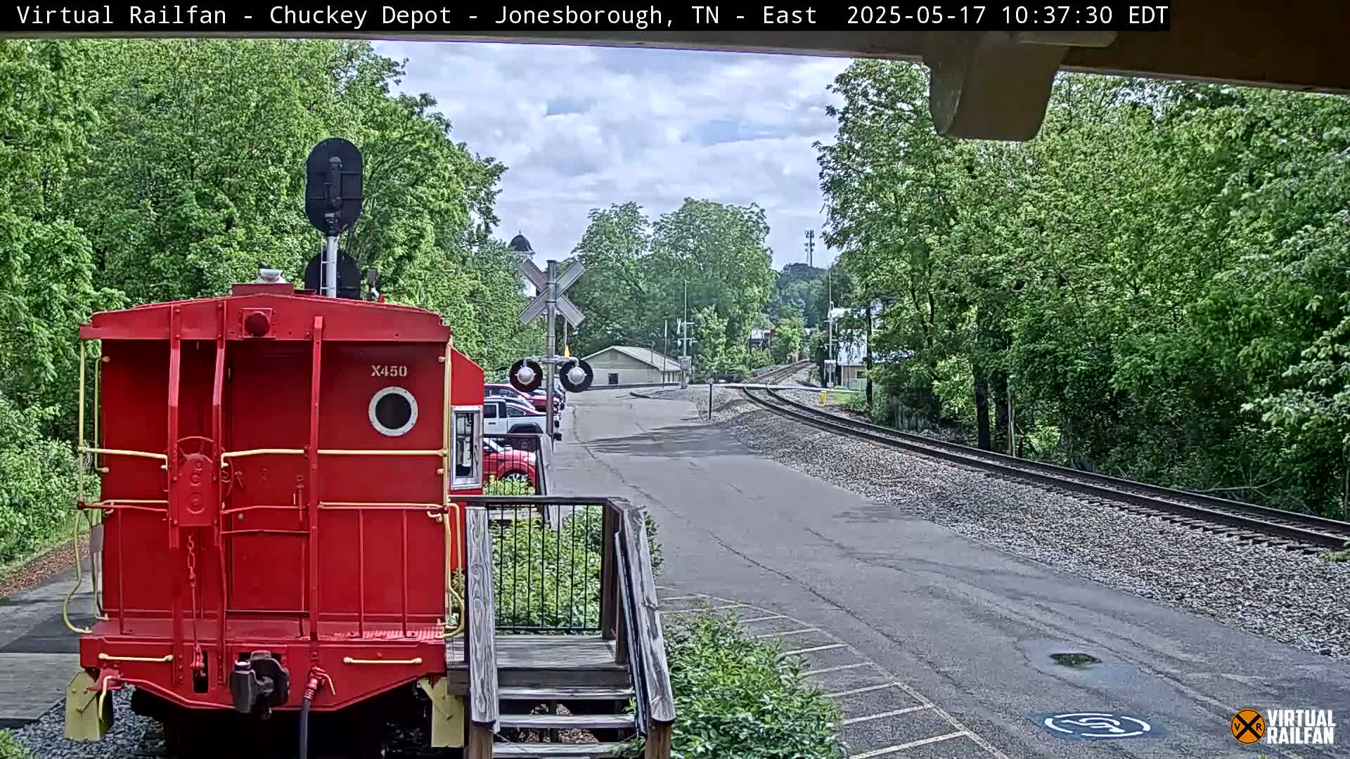 A red railroad car is parked facing away from the camera at a train depot on a partly cloudy day.
