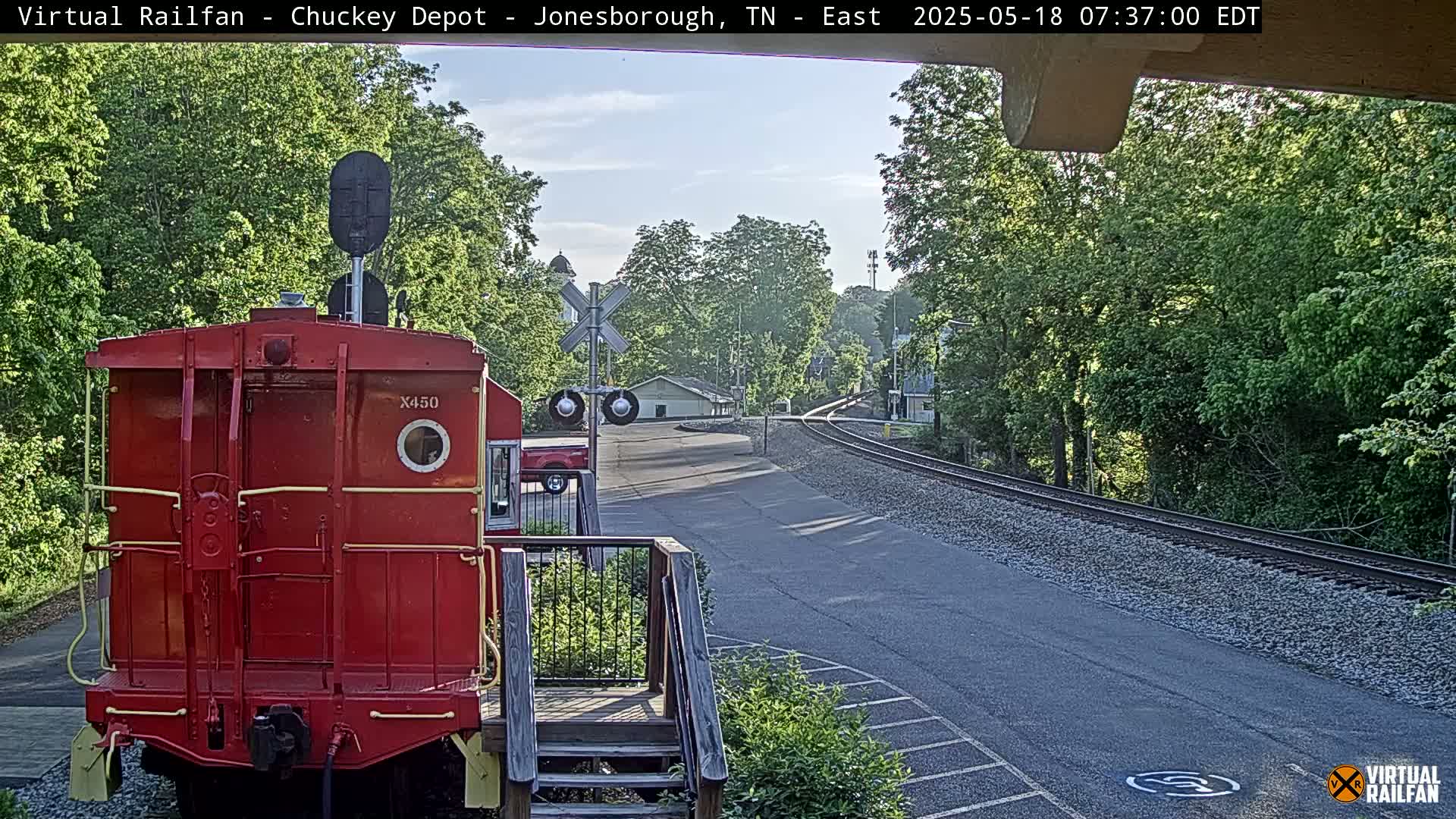 A red railroad car is parked at a train depot on a sunny morning, with a railroad track and lush green trees in the background.