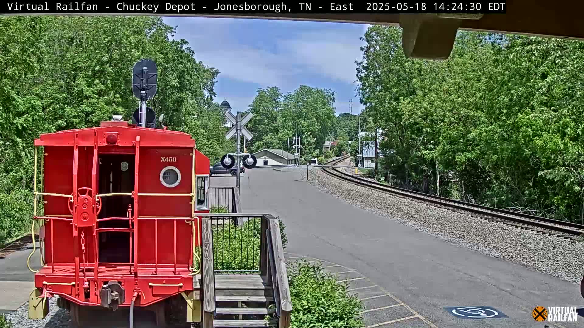 A red railroad car sits at a train depot on a sunny day, with train tracks curving into a wooded area.