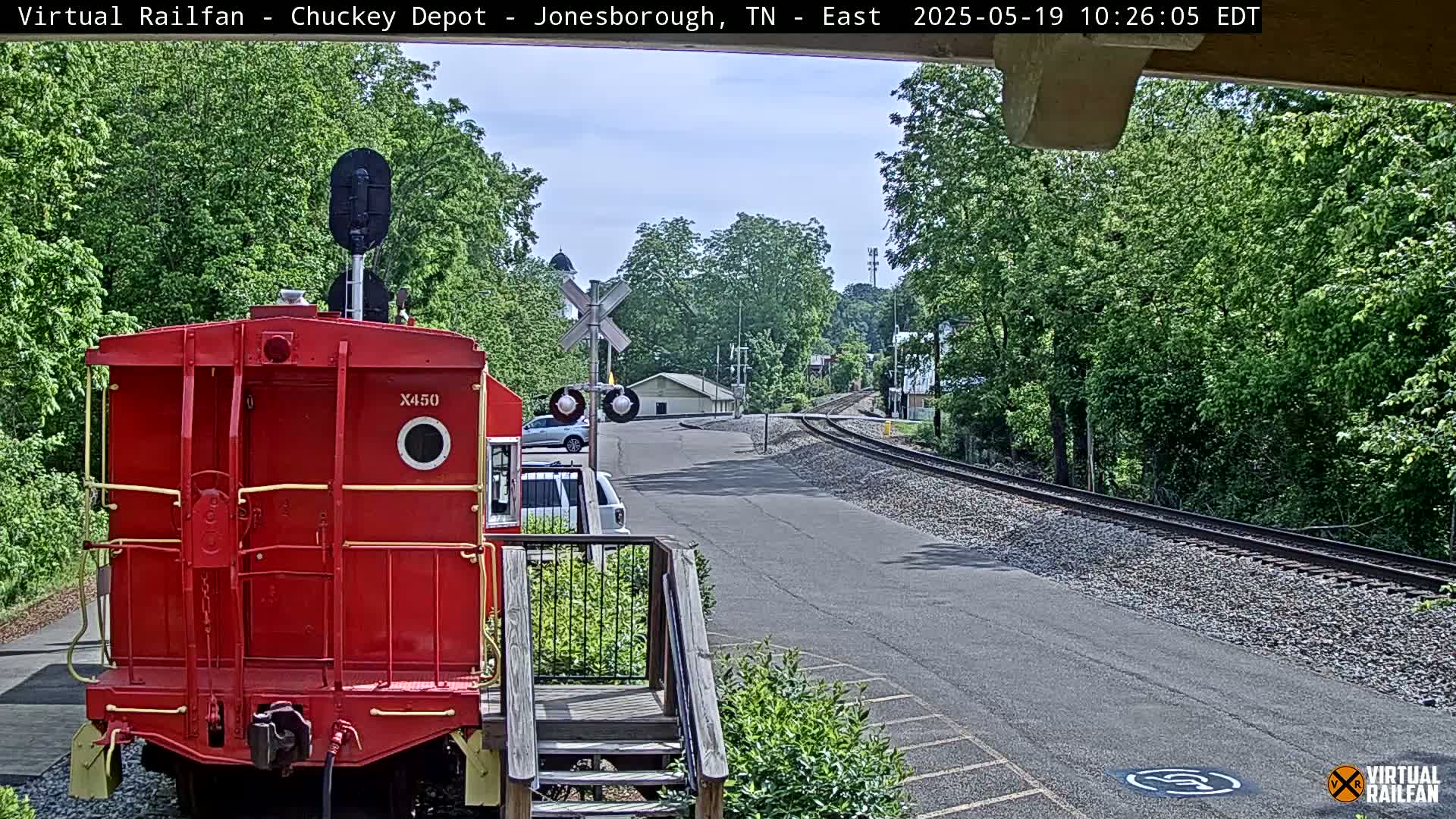 A red railroad car sits facing away from a paved road and railroad tracks that curve away into a green, leafy landscape under a mostly clear sky.