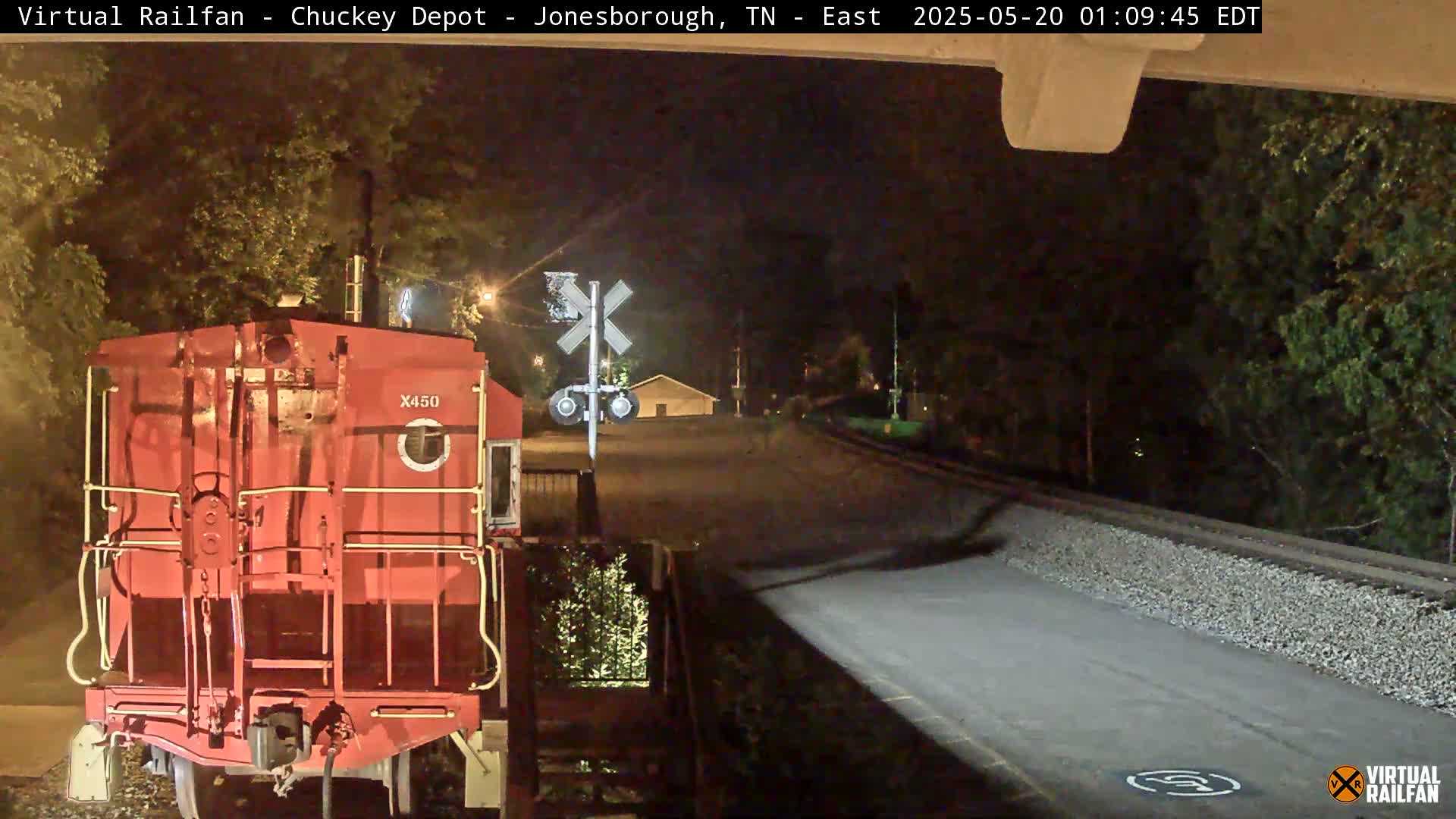 At night, the rear of a red train car is visible near a railroad crossing, with trees and a road in the background.