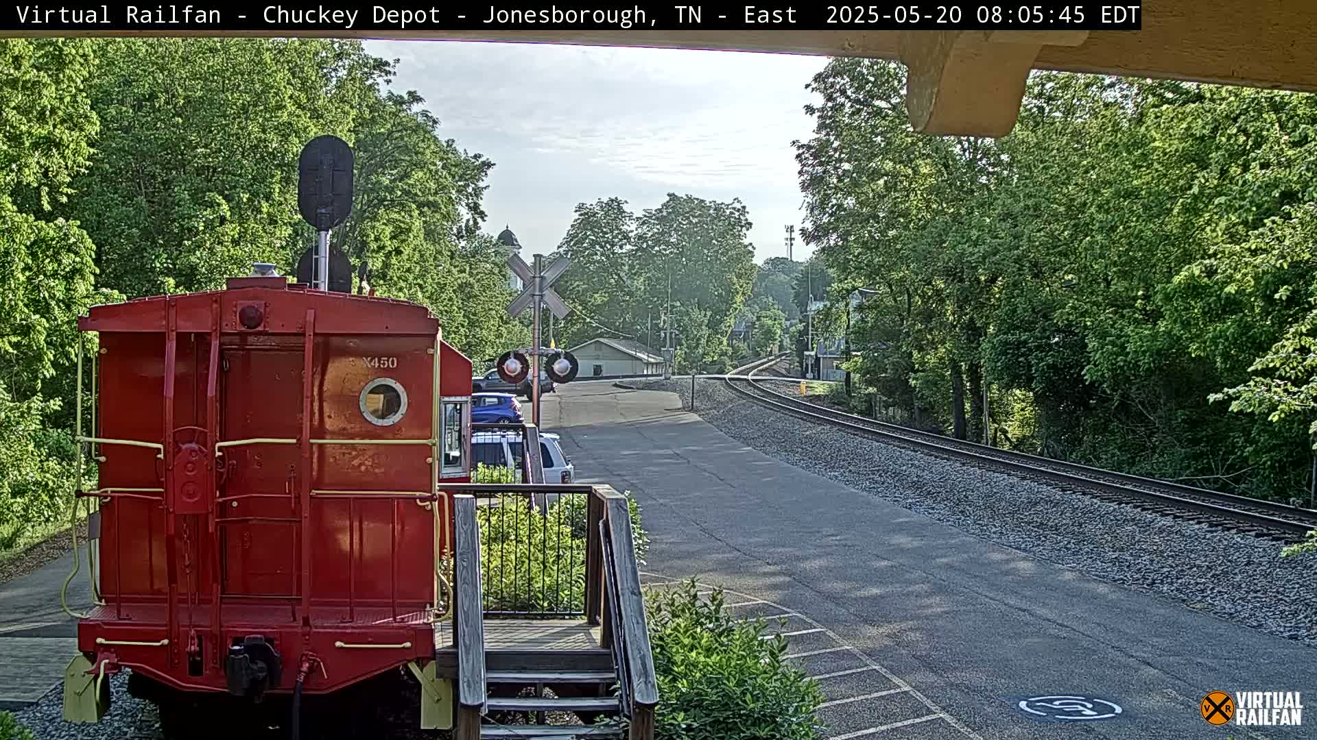 A red railroad car sits in front of a railway line curving into a tree-lined town under a partly sunny sky.