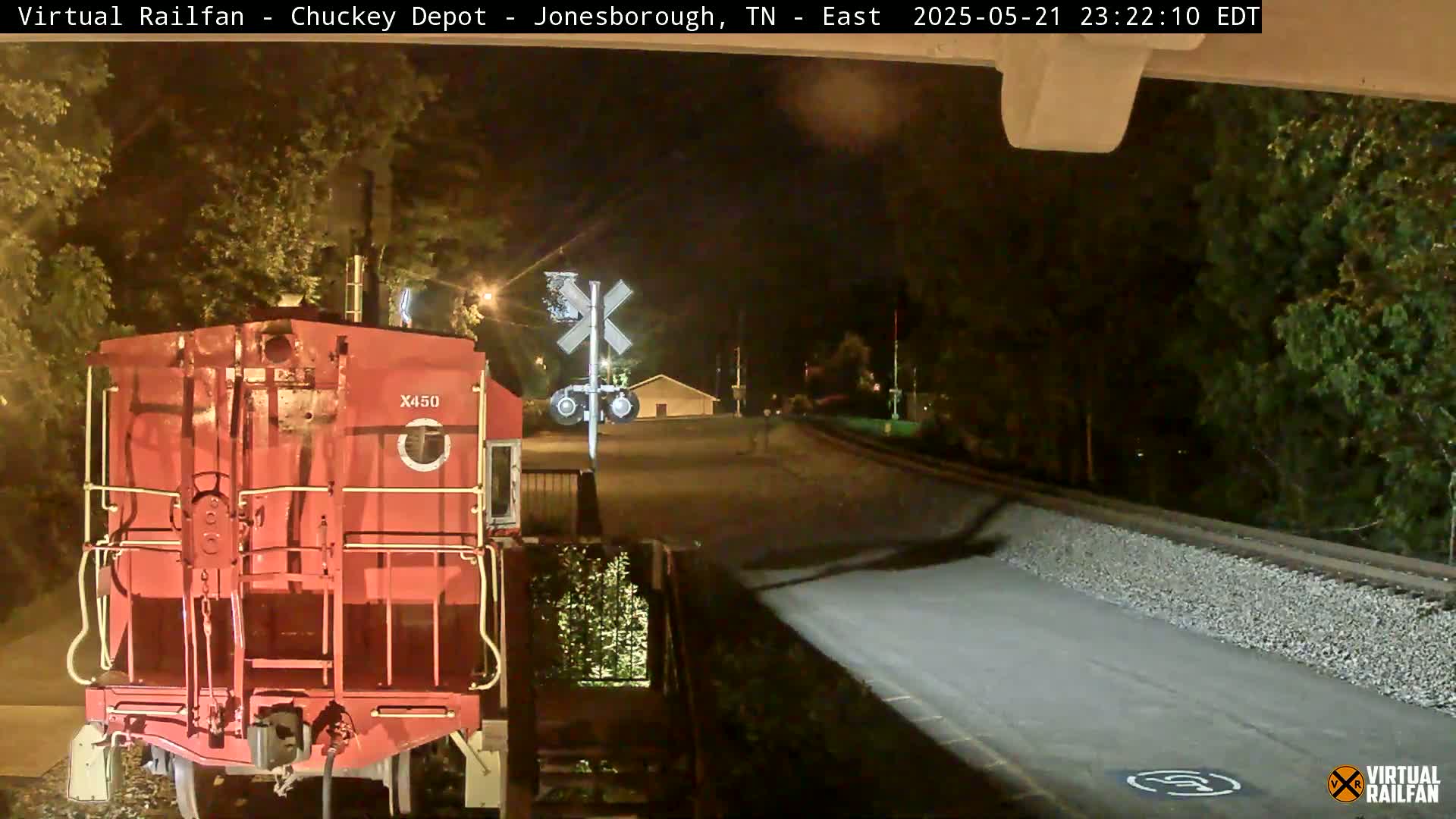 At night, a red train car sits at a railroad crossing near a road, with trees and a building visible in the background.
