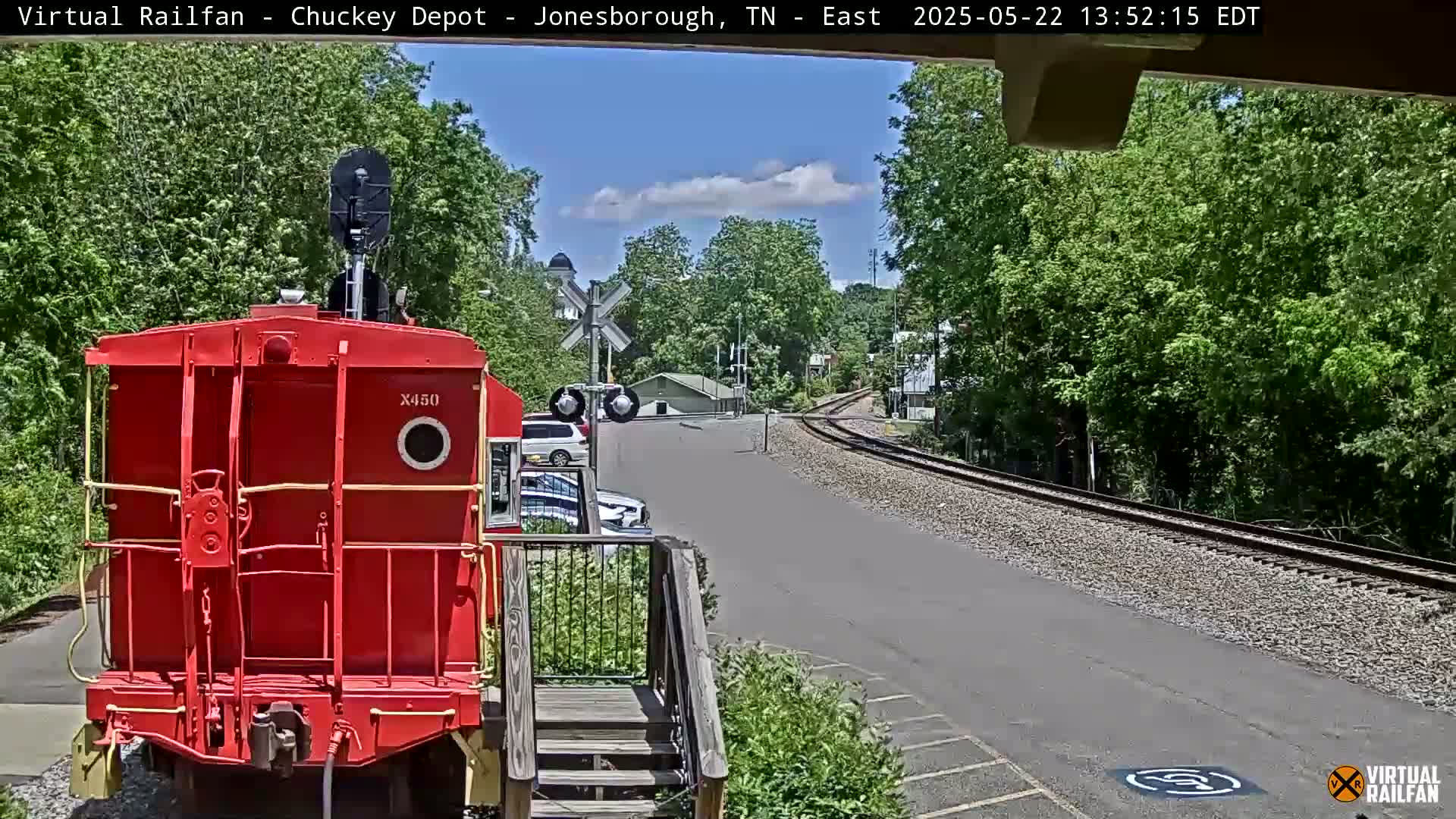 A red railway car is parked facing the camera, with railroad tracks and a partly sunny sky visible beyond it.