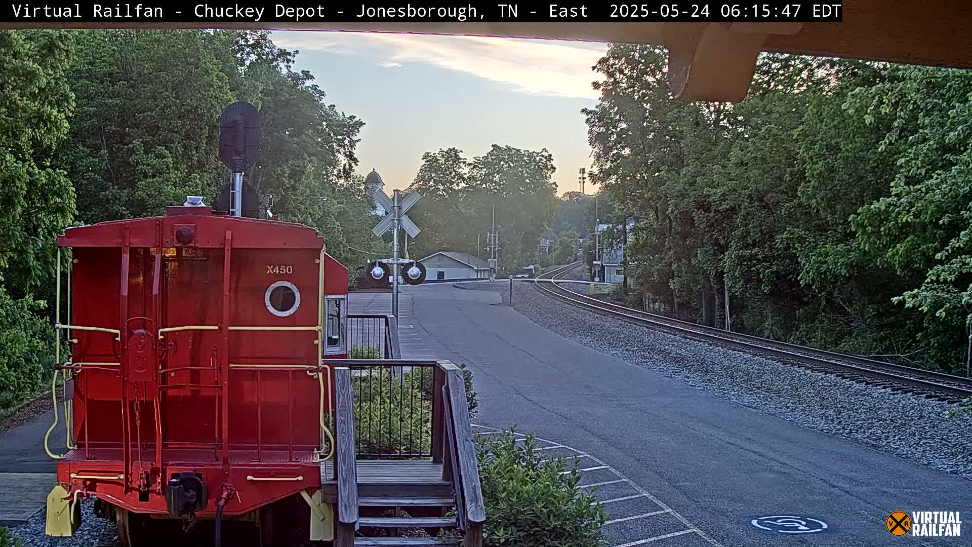 A red railroad car is parked facing away from the camera, overlooking a railroad track curving into a town under a twilight sky.