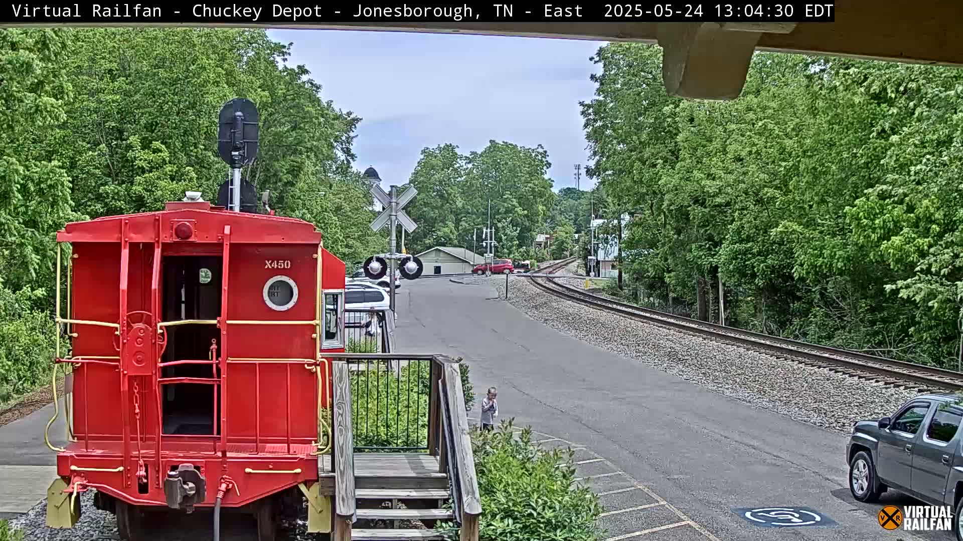 A red train caboose is parked at a train depot on a sunny day, with a child walking nearby and train tracks curving into the distance.