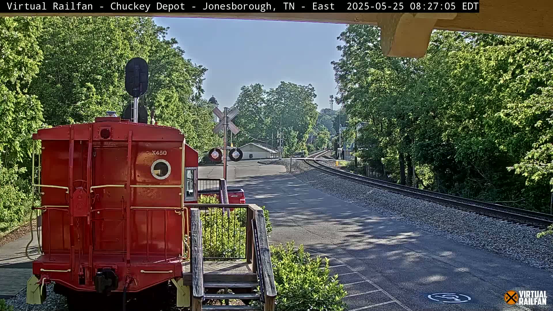 A red railroad car is parked at a train depot on a sunny day, with railroad tracks extending into a tree-lined area.